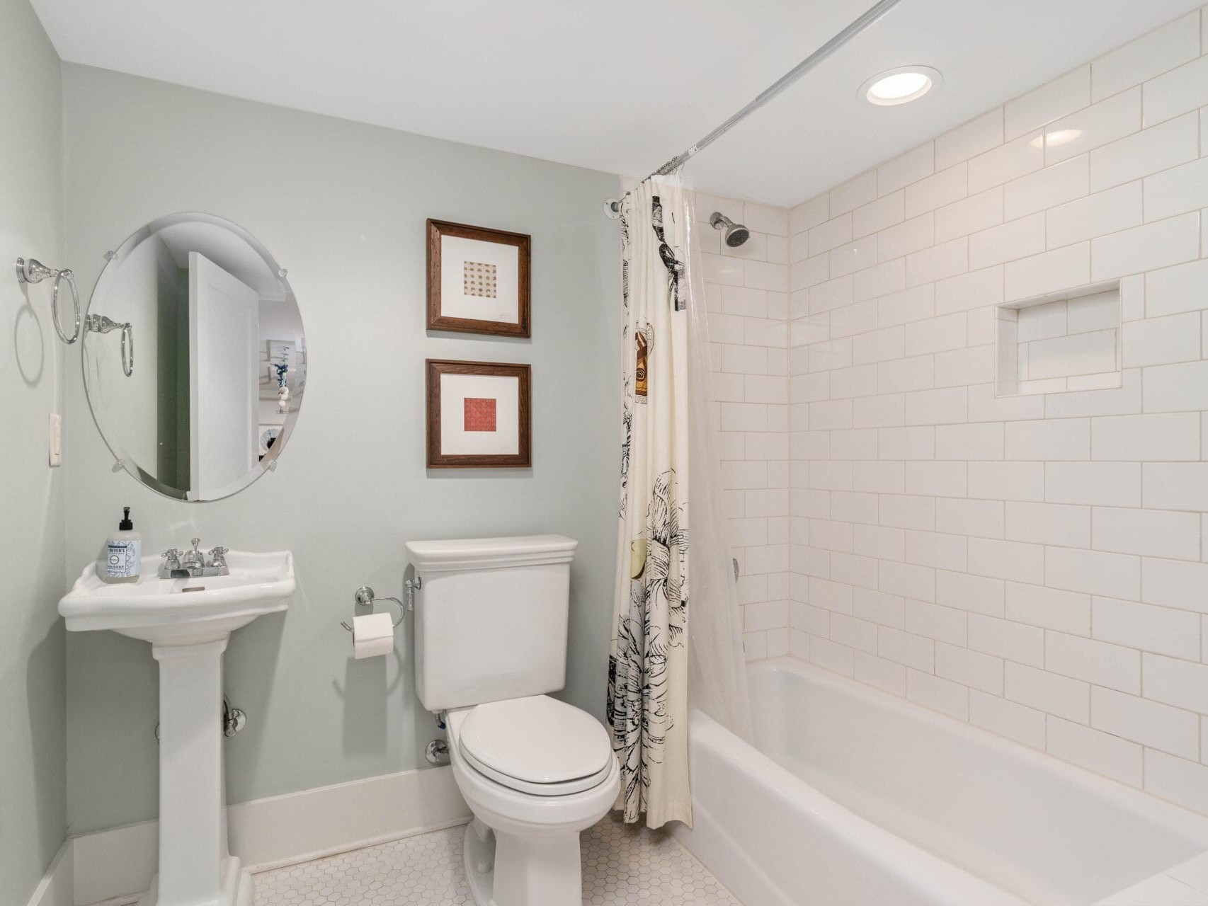 A bathroom featuring a white toilet and pedestal sink on the left. Theres a white bathtub with a shower curtain on the right, and two framed pictures hang on the light green wall above the toilet. The floor is covered with small white tiles.