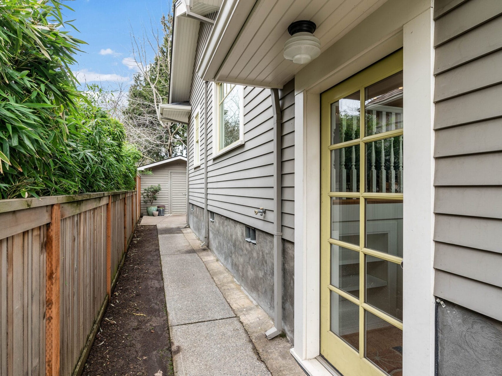 Side walkway of a house with gray siding, featuring a door with glass panels. A wooden fence borders the path on the left, and bamboo plants add greenery. A small structure is visible at the end of the path. The sky is partly cloudy.