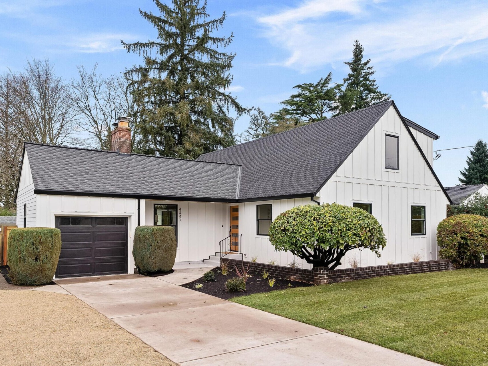A modern white house with a dark roof stands proudly, featuring a single garage door. The front yard boasts a well-maintained lawn, shrubs, and a paved driveway leading to the entrance. Endorsed by top Portland real estate agents, tall trees and a bright blue sky form the perfect backdrop.