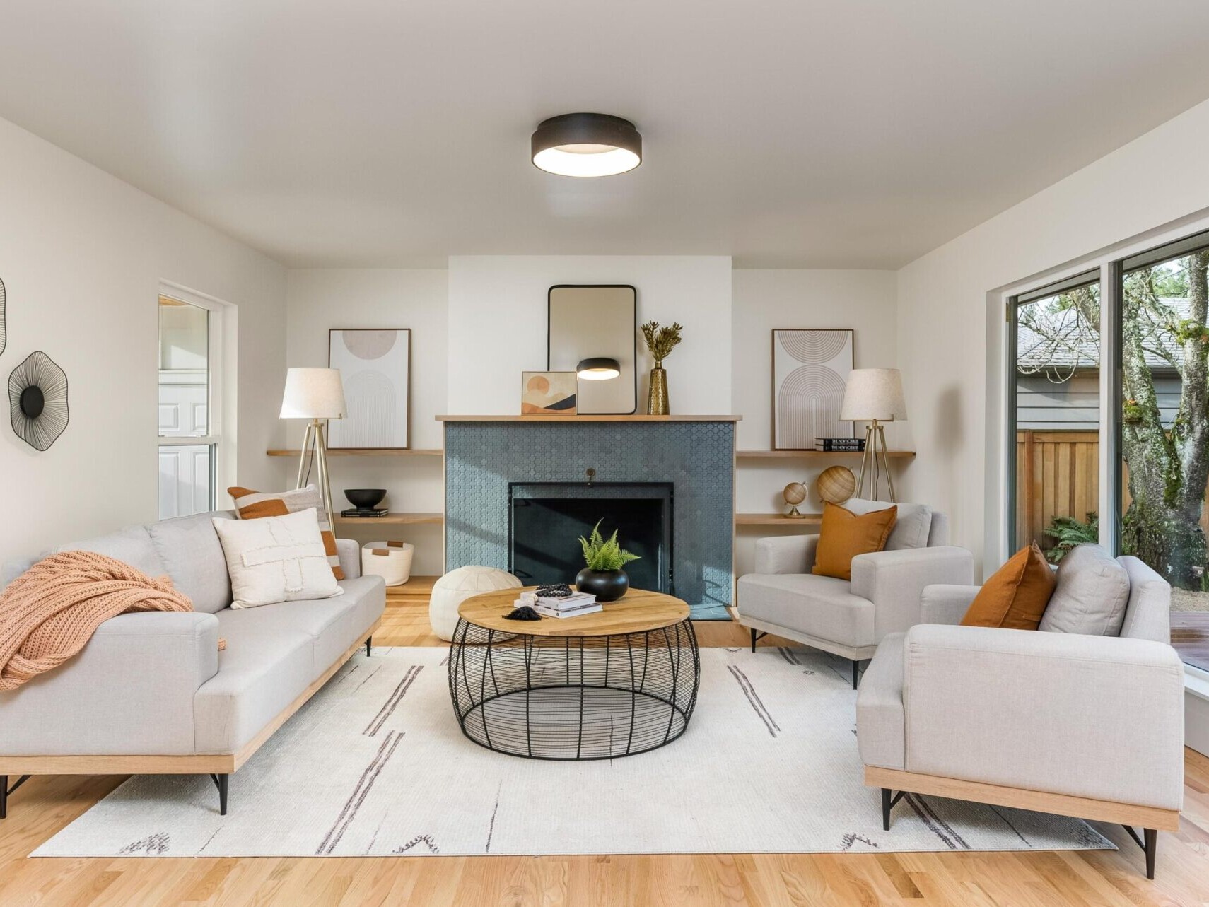 A modern living room in Portland Oregon real estate with beige sofas, a round coffee table, and a fireplace. The space features minimalist decor, wall art, a potted plant, and neutral tones. A large window on the right lets in natural light while wooden floors add warmth.
