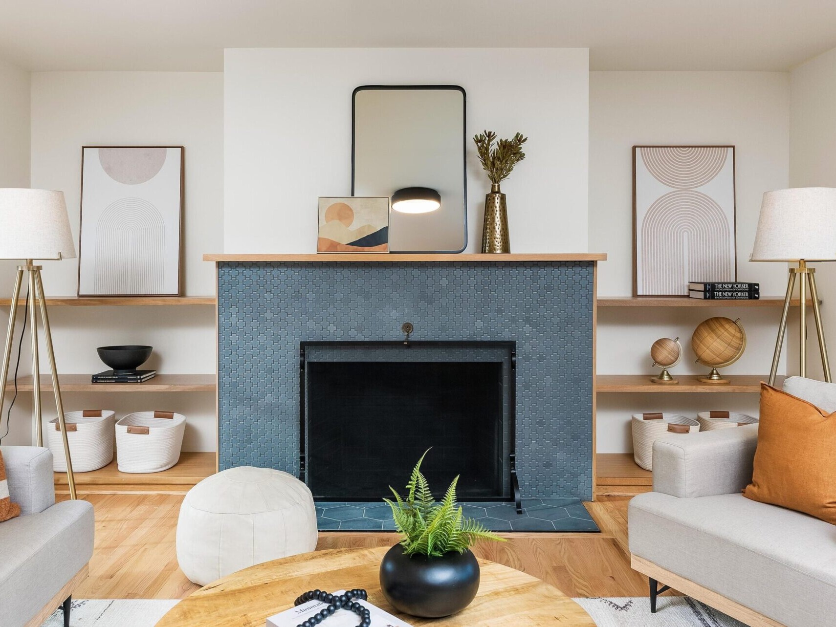 A modern living room with a minimalist design in a Portland real estate gem. Features include a black tiled fireplace, abstract artwork, a round mirror, and decor items like books and vases, all in neutral tones. Two beige sofas and a wooden coffee table complete the look.