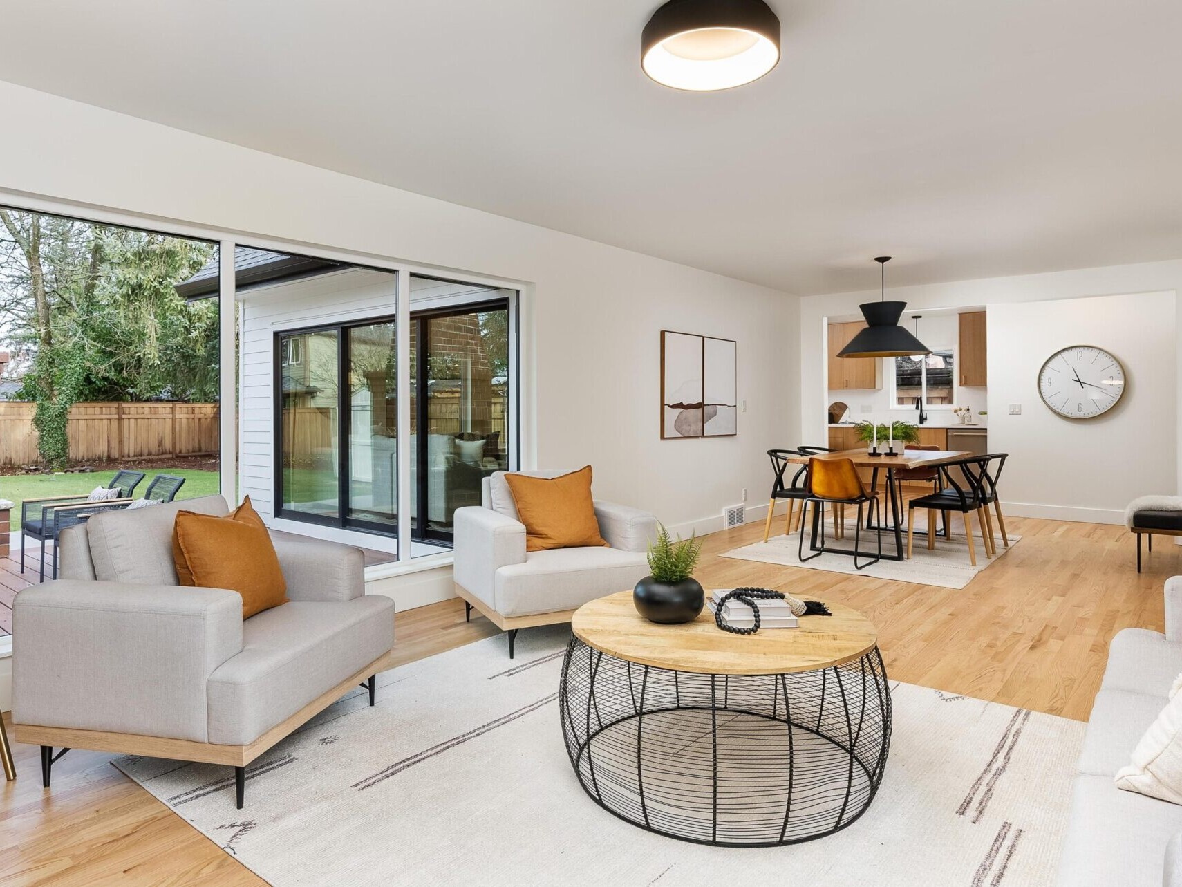 A modern living room with light wood flooring and white walls. It features two gray armchairs, a circular wireframe coffee table, and a gray sofa with brown cushions. Discover the elegance of Portland Oregon real estate, highlighted by a dining area near large sliding glass doors.