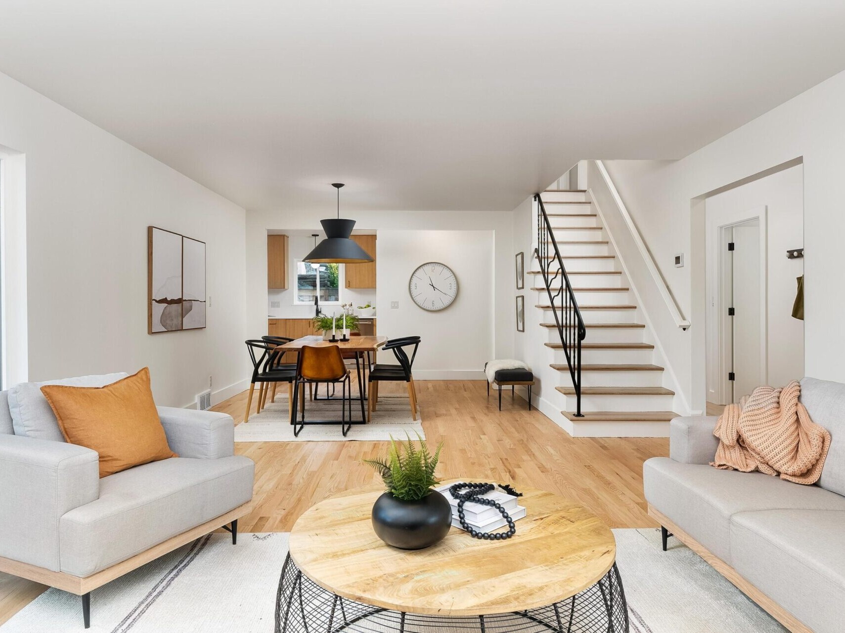A modern living room in a Portland Oregon real estate gem features light gray sofas, orange cushions, and a round wooden coffee table. A dining area with table and chairs graces the background, while wooden flooring and stairs with a black railing complete this neutral-toned haven.