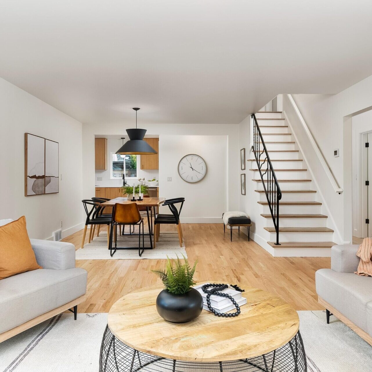 Modern living room with light wood floors, a round coffee table, and two gray sofas accented by orange cushions. In the background, a dining area features a rectangular table and black chairs, alongside a staircase leading upstairs. A large clock adorns the wall in this stylish Portland real estate gem.