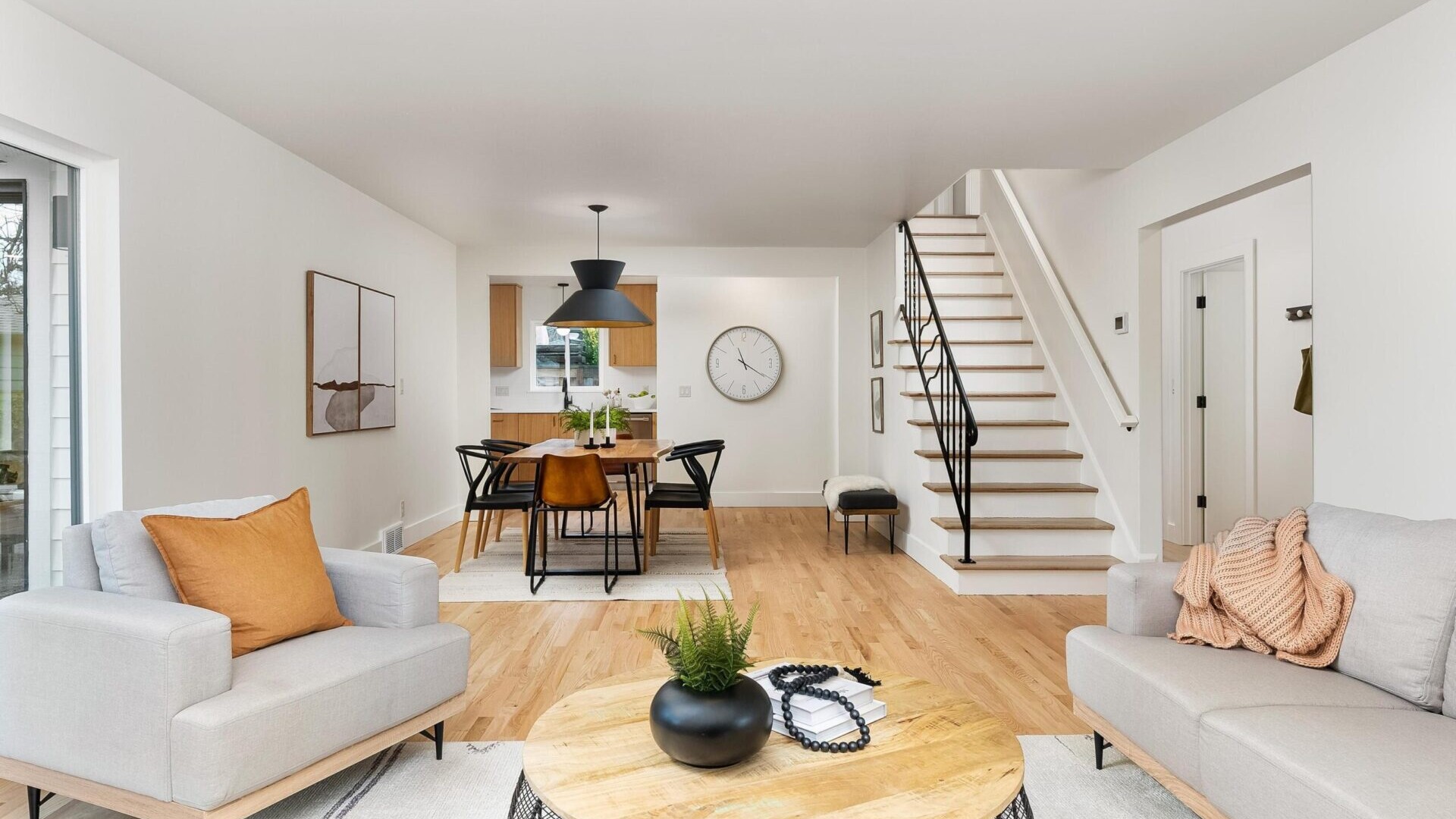 Modern living room with light wood floors, a round coffee table, and two gray sofas accented by orange cushions. In the background, a dining area features a rectangular table and black chairs, alongside a staircase leading upstairs. A large clock adorns the wall in this stylish Portland real estate gem.
