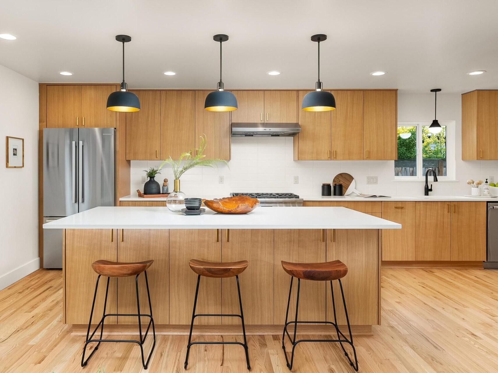 A modern kitchen in Portland real estate showcases wooden cabinetry and a large white island. Black pendant lights hang above, accompanied by three wooden bar stools. Stainless steel appliances and a decorative bowl complete this elegant space, highlighted by a skilled Portland realtor.