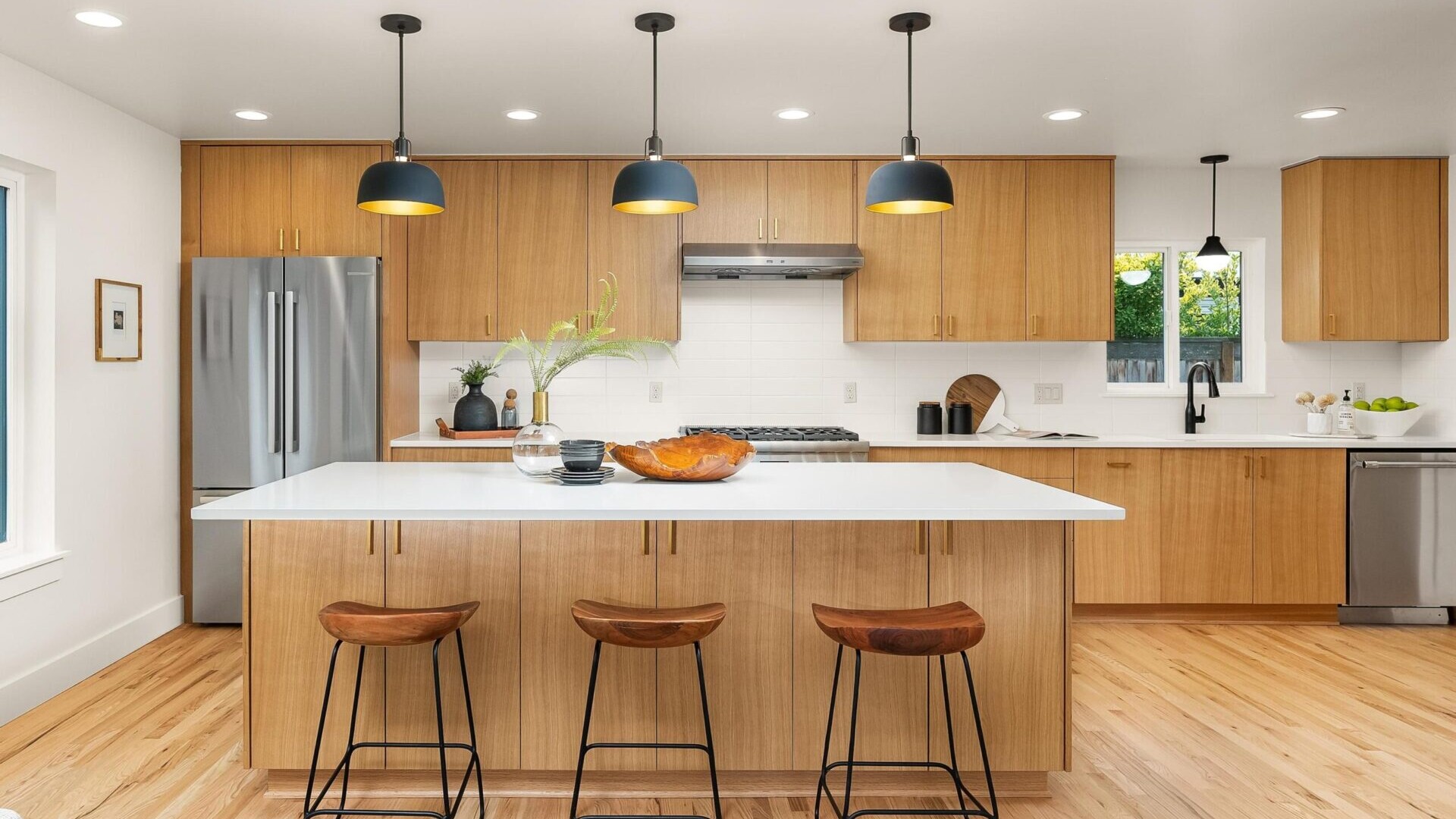 Modern kitchen with light wood cabinetry, a central island featuring a white countertop, and three wooden barstools. Overhead, black pendant lights delicately illuminate the space. Stainless steel appliances and decorative items complete this warm, inviting jewel in Portland real estate.