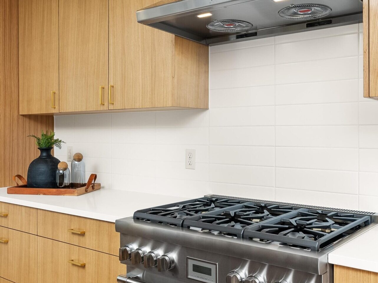 A modern kitchen in a Portland Oregon real estate gem features wooden cabinets and a stainless steel oven. A sleek range hood sits above the stove with a white tile backsplash. A black teapot and wooden utensils adorn the white countertop, showcasing meticulous attention to detail by your top real estate agent.