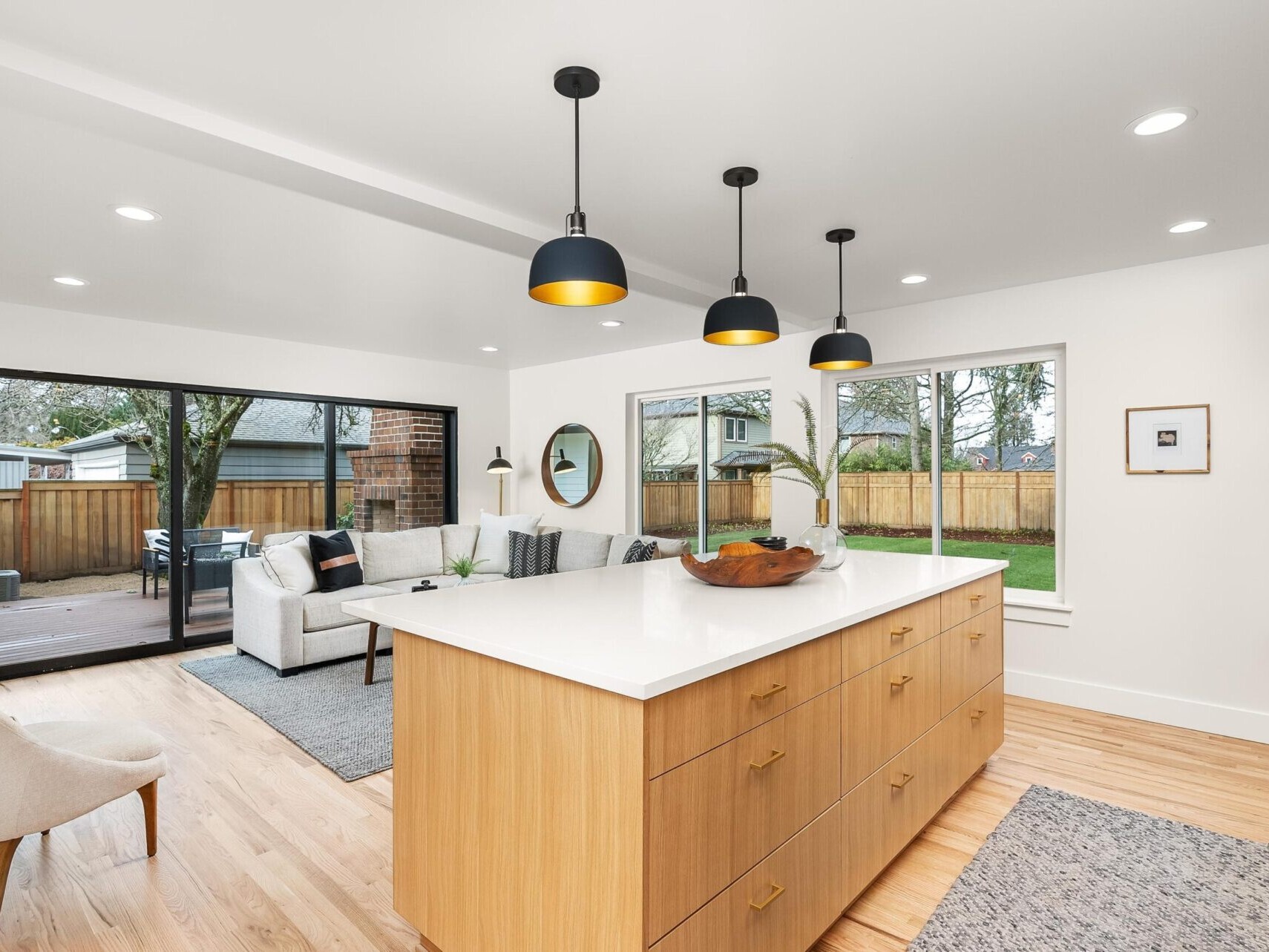 Modern kitchen and living area with a large wooden island, pendant lights, and a sofa. Glass sliding doors open to a patio with outdoor furniture. Light wood floors and white walls create a spacious feel. Recommended by Portland Oregon Real Estate experts, the large windows provide natural light.
