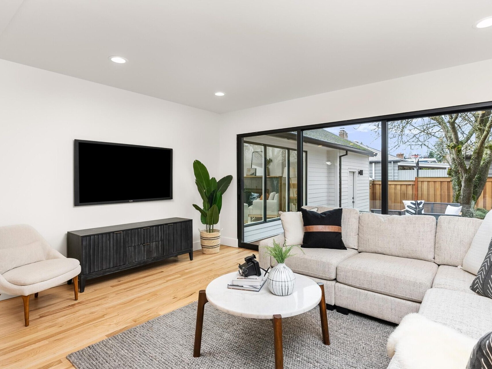 A modern living room in a Portland Oregon real estate gem features a beige sectional sofa, round marble coffee table, and potted plant. A wall-mounted TV and large window overlooking the backyard enhance the space, completed by a light gray armchair and dark sideboard.