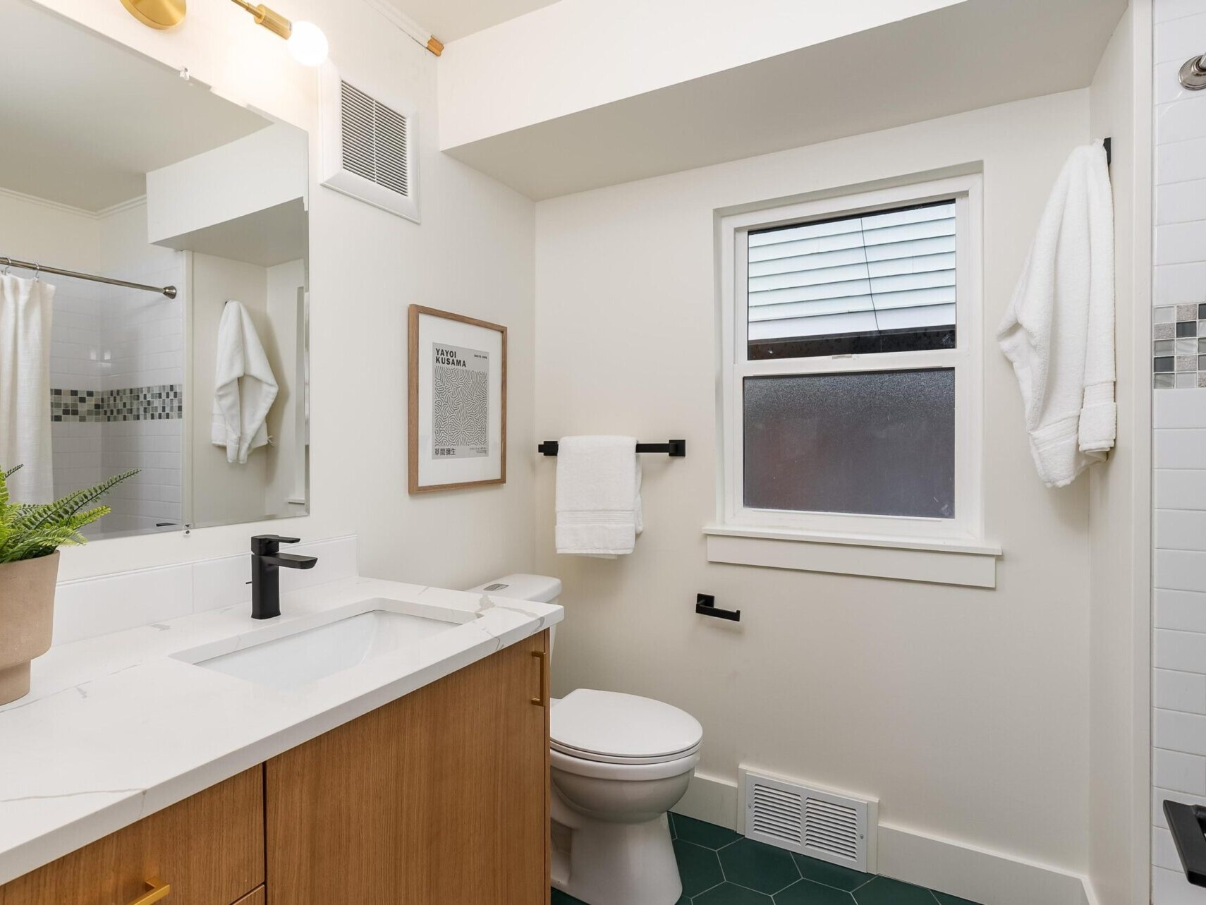 A modern bathroom in a Portland Oregon real estate gem features a wooden vanity, white countertop, and sleek black fixtures. A potted plant adds a touch of nature. The white tiled shower boasts a black and white mosaic border, while natural light streams through the window above the toilet.