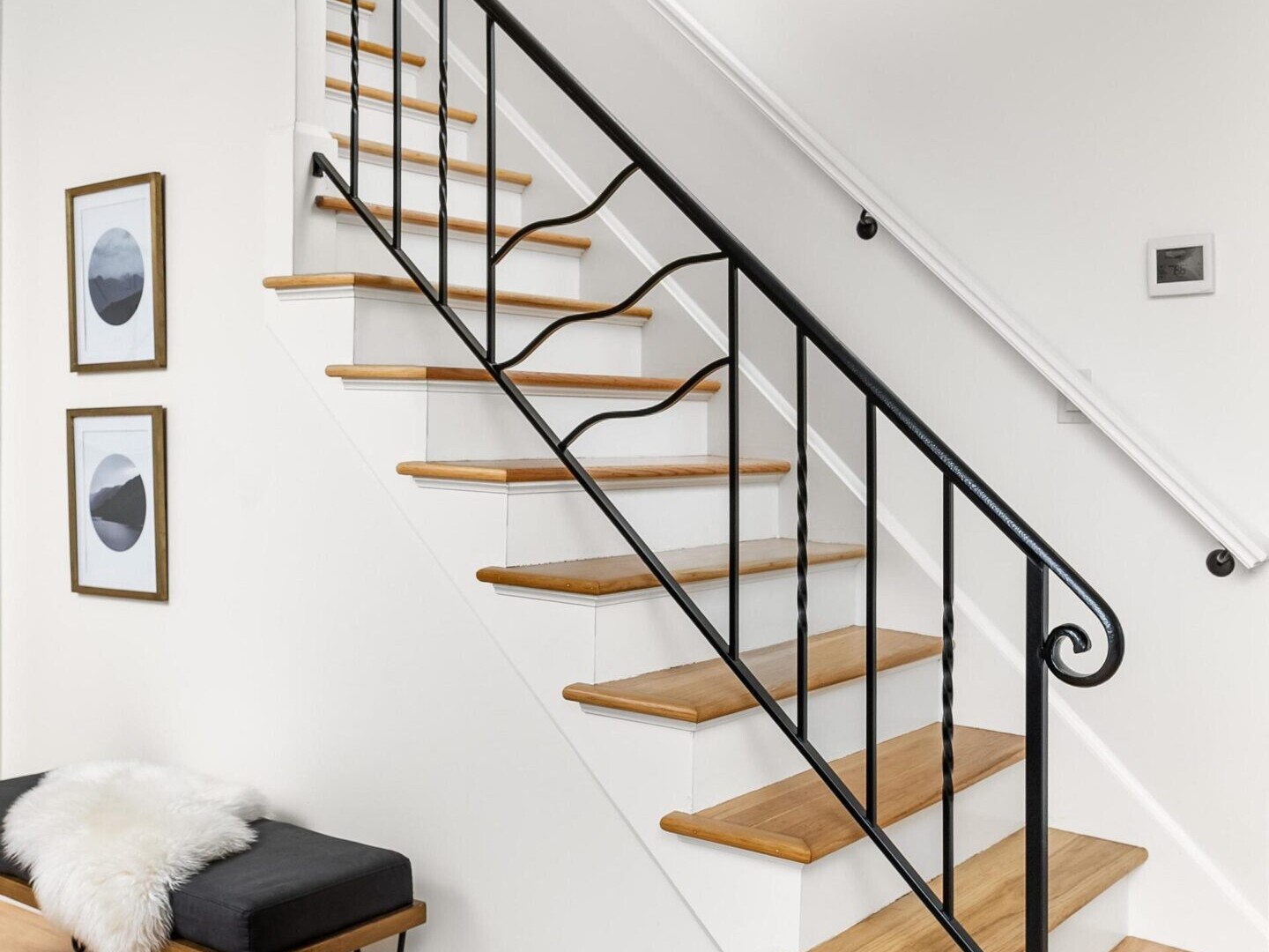 A modern staircase with light wood steps and a black metal railing showcases the elegance often highlighted by a top Portland Realtor. Below, a black bench with metal legs is adorned with a white fluffy cushion. Two framed abstract prints hang on the white wall beside the stairs.