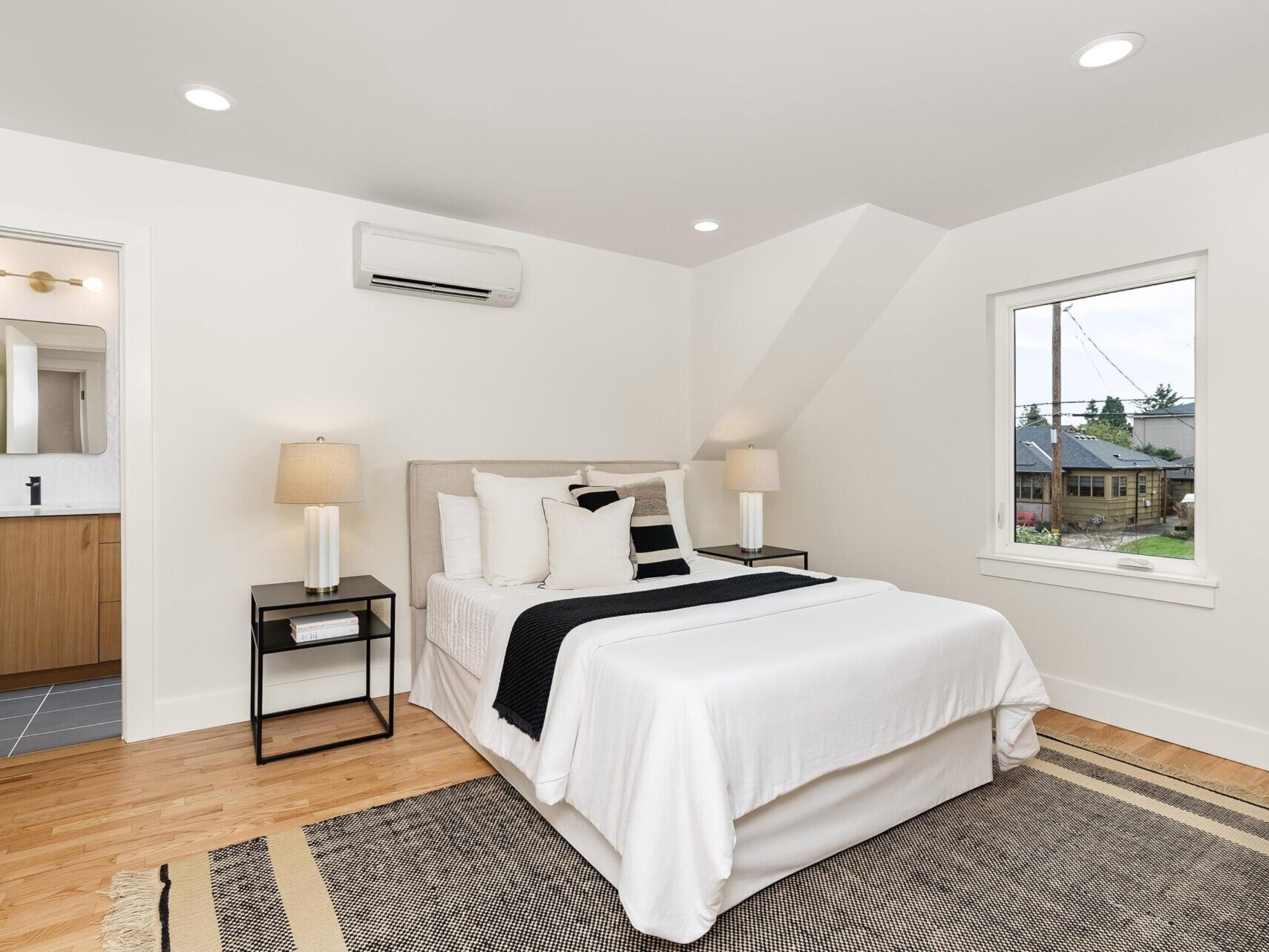 A modern bedroom in Portland real estate features a neatly made bed with white and black bedding. Two bedside tables with lamps flank it, while a rug adorns the wooden floor. An open door offers a glimpse of the bathroom, and the window provides a street view.
