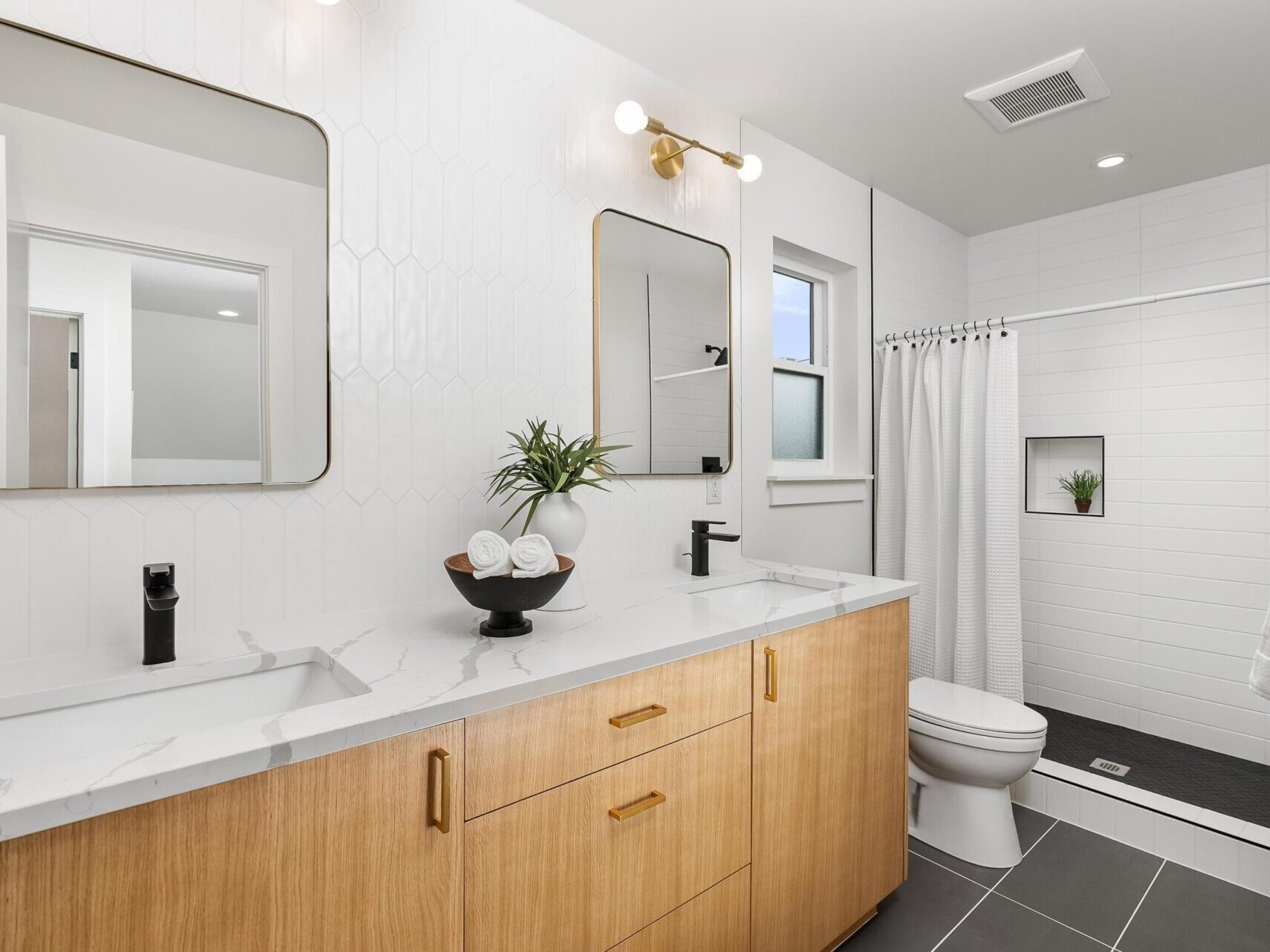 A modern bathroom with a white marble countertop featuring dual sinks, wooden cabinetry, and black fixtures complements this exquisite Portland Oregon real estate listing. Two mirrors sit above the sinks while a glass-enclosed shower graces the right, accented by white tiles and a small potted plant.