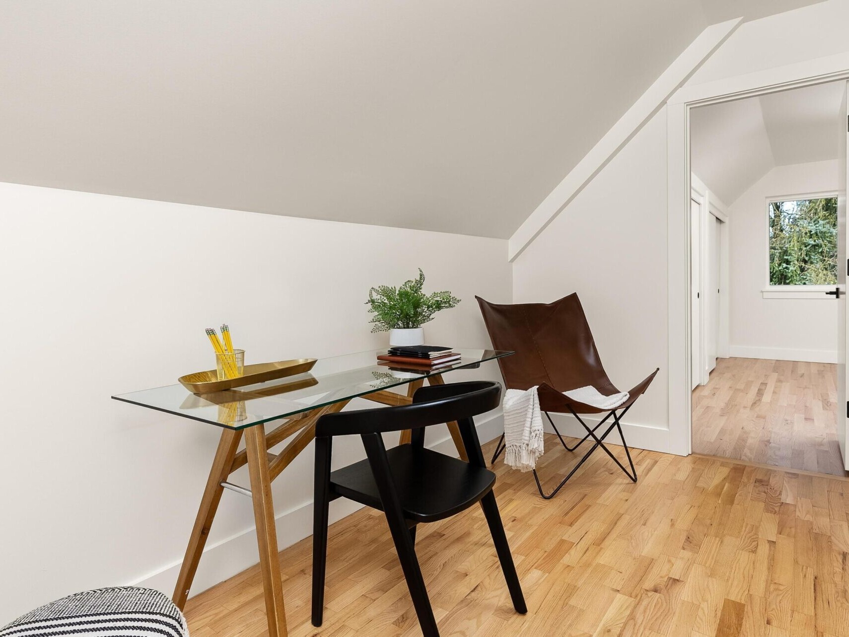 Minimalist attic office in Portland Realtor style with a glass desk, black chair, and leather butterfly chair. A potted plant, books, and pencils adorn the desk. Light wooden floor and sloped white ceiling enhance the bright, airy feel. Doorway leads to another room.