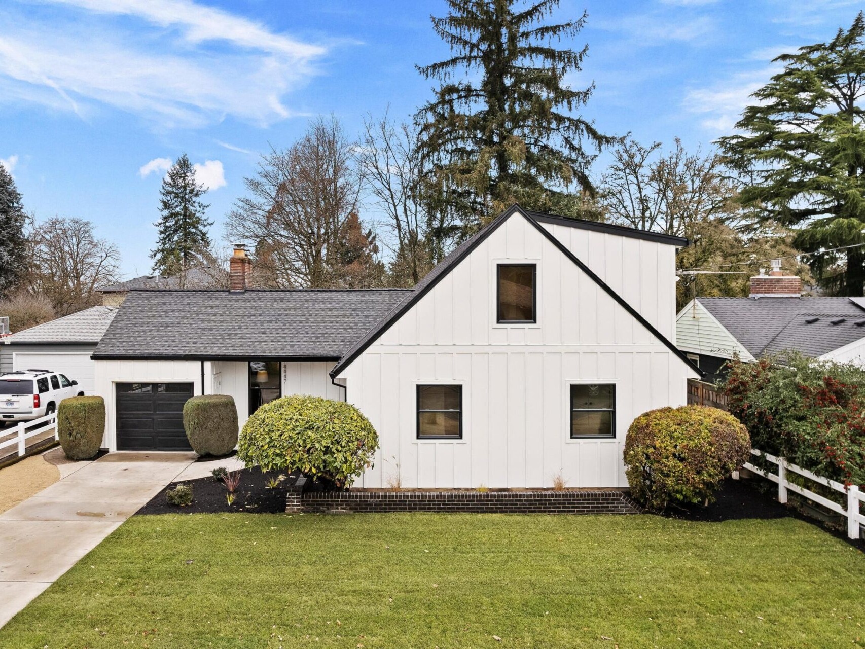 A modern white house with a black roof, featuring an attached garage and a well-maintained front yard, is listed by the top Portland Realtor. Surrounded by a white picket fence and lush greenery, large trees are visible in the background under a partly cloudy sky.