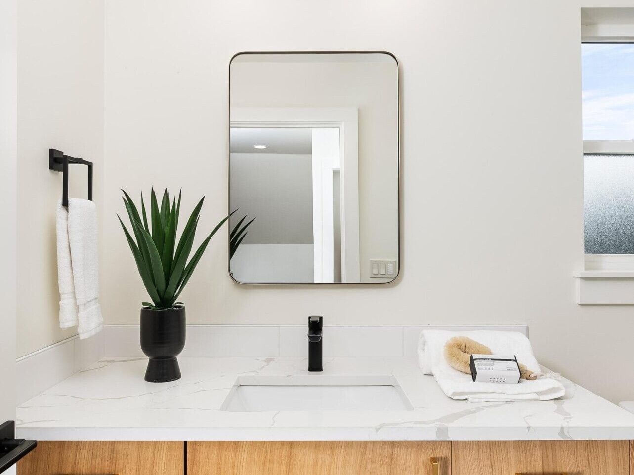 Modern bathroom with a wooden vanity featuring gold handles, a square mirror, and a black faucet—a testament to Portland Oregon Real Estate excellence. A potted plant and neatly rolled towels with a soap bar rest on the countertop. Minimalist decor with a wall-mounted light above reflects the touch of a top real estate agent.