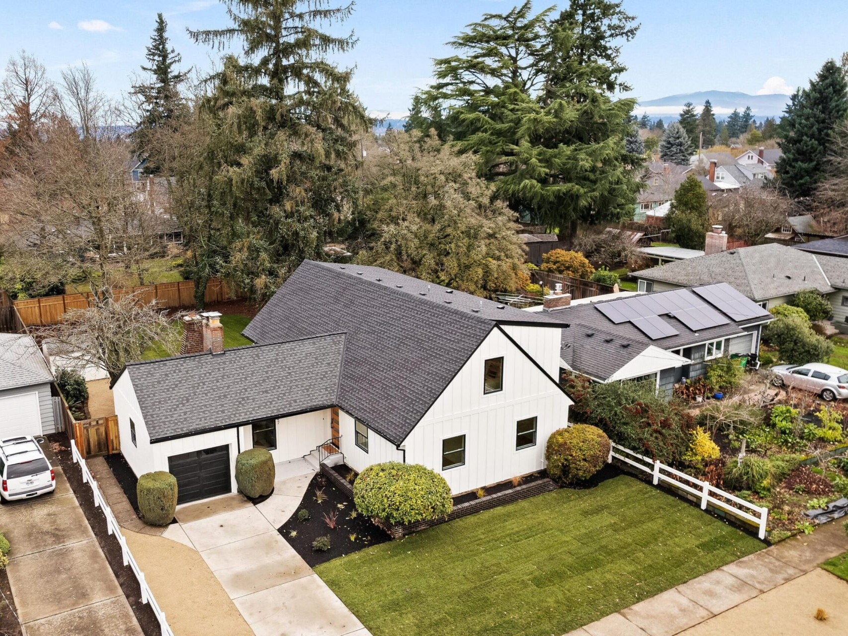 Aerial view of a white two-story house with a dark roof and garage in Portland. It features a well-maintained lawn, a white fence, and solar panels on the neighboring house. Tall trees and other houses surround the property under a partly cloudy sky, showcasing Portland Oregon real estate charm.