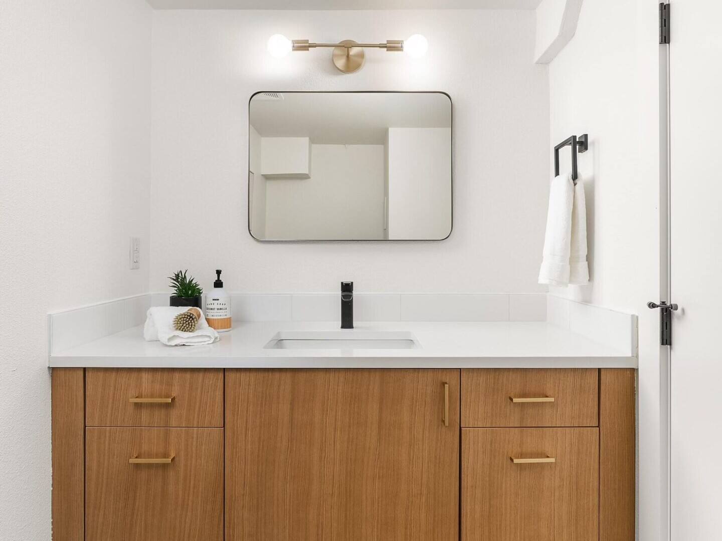 A modern bathroom exudes style with a wooden vanity paired with a white countertop, black faucet, and rectangular mirror. The white wall hosts two round lights above the mirror. A towel and soap dispenser sit on the counter. Perfect details for showcasing in Portland Oregon real estate listings.