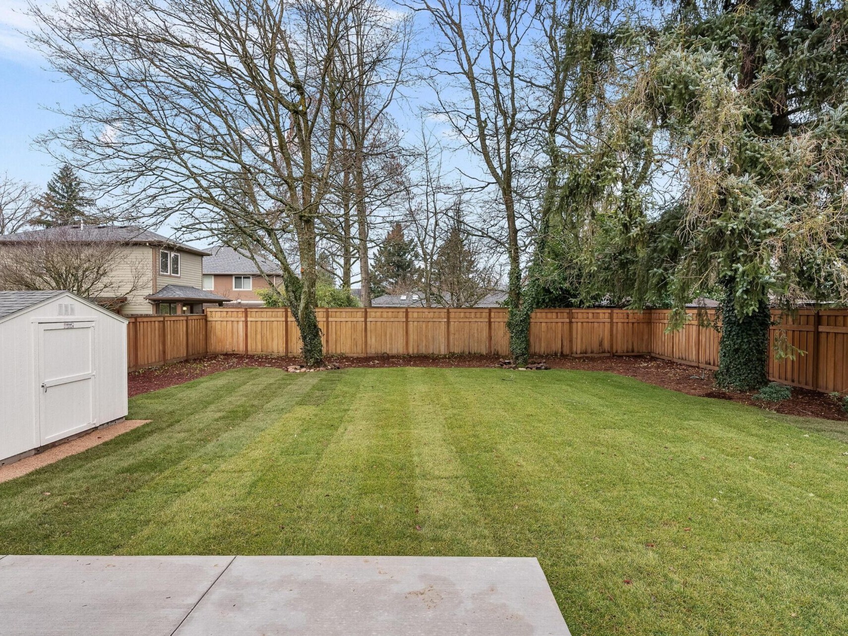 A manicured backyard with freshly mowed grass, surrounded by a wooden fence, offers a serene escape. Tall trees with sparse leaves line the perimeter. A white shed sits on the left under a partly cloudy sky. Discover such peaceful retreats with a top real estate agent in Portland, Oregon.