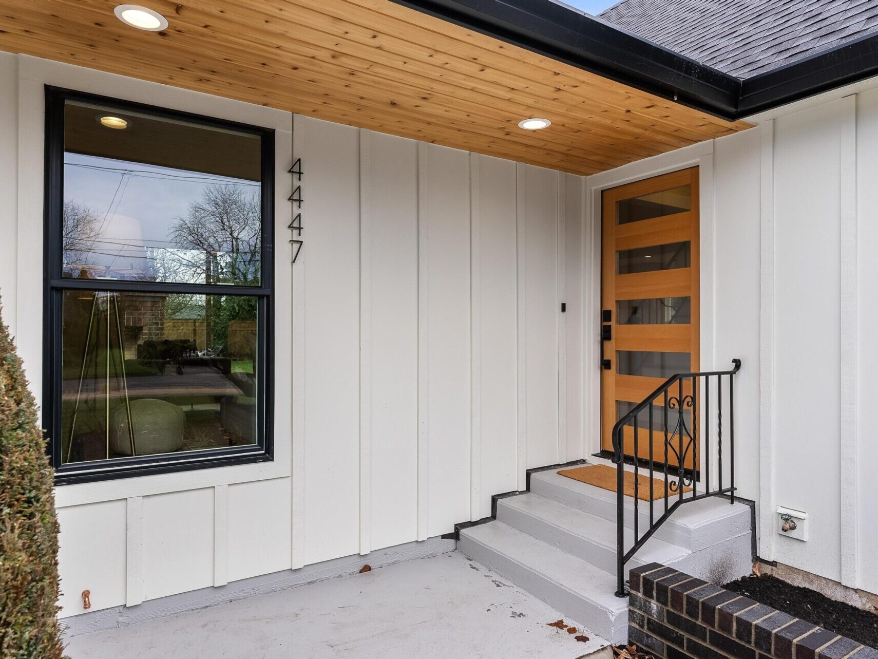  A modern house entrance in Portland, Oregon, features a wooden door with a horizontal glass panel design. The white siding and black-framed window create a sleek look. Steps with a black railing lead to the door, and a small hedge is visible on the left—a true gem in Portland real estate.