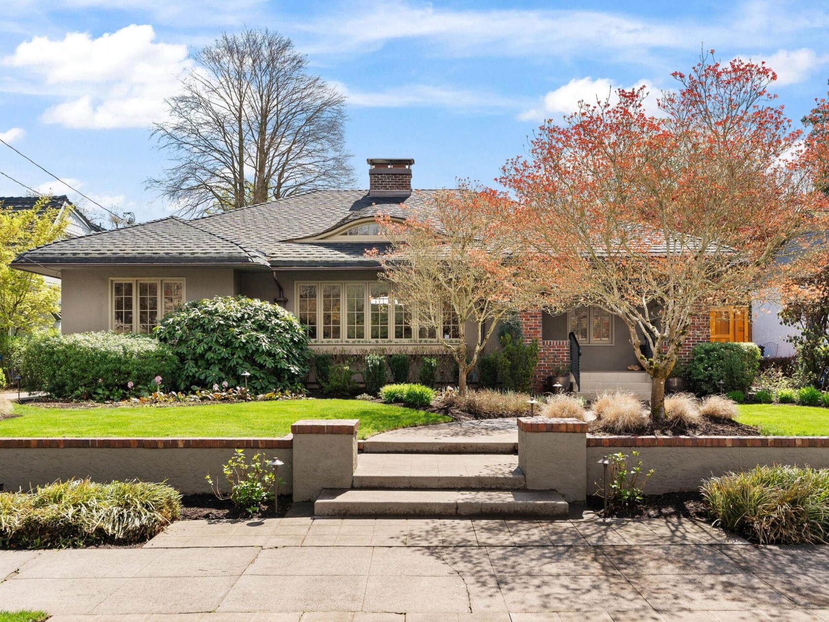 Single-story house with a gray roof and beige walls, surrounded by a well-maintained garden with trees and bushes. The walkway leads to steps up to the front entrance. A clear blue sky is overhead—a perfect gem for anyone exploring Portland Oregon real estate.