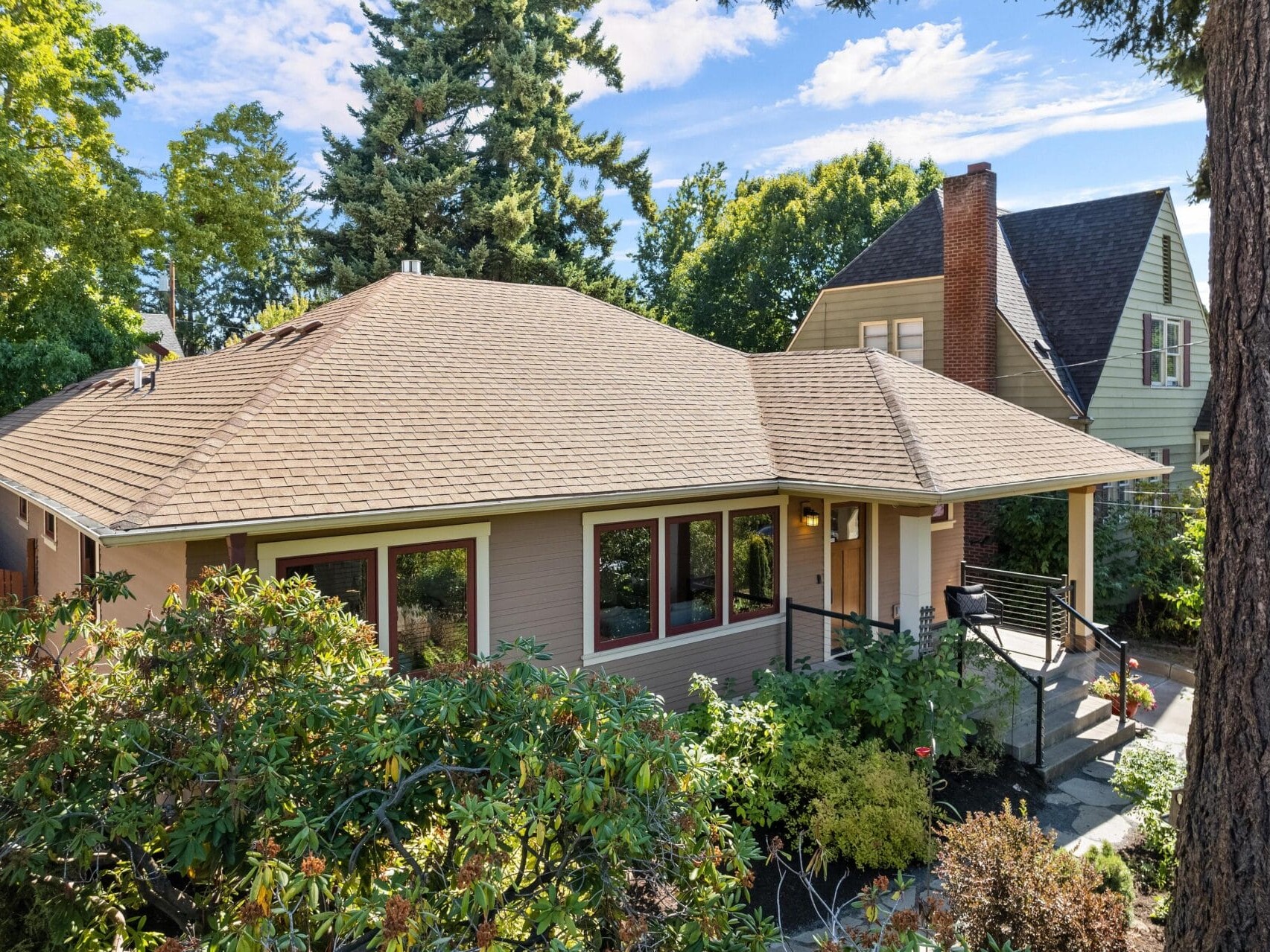 A charming single-story house in Portland, Oregon, features a brown roof surrounded by lush green trees and shrubs. A chimney rises on the right, and a path leads to the entrance, all under a bright blue sky.