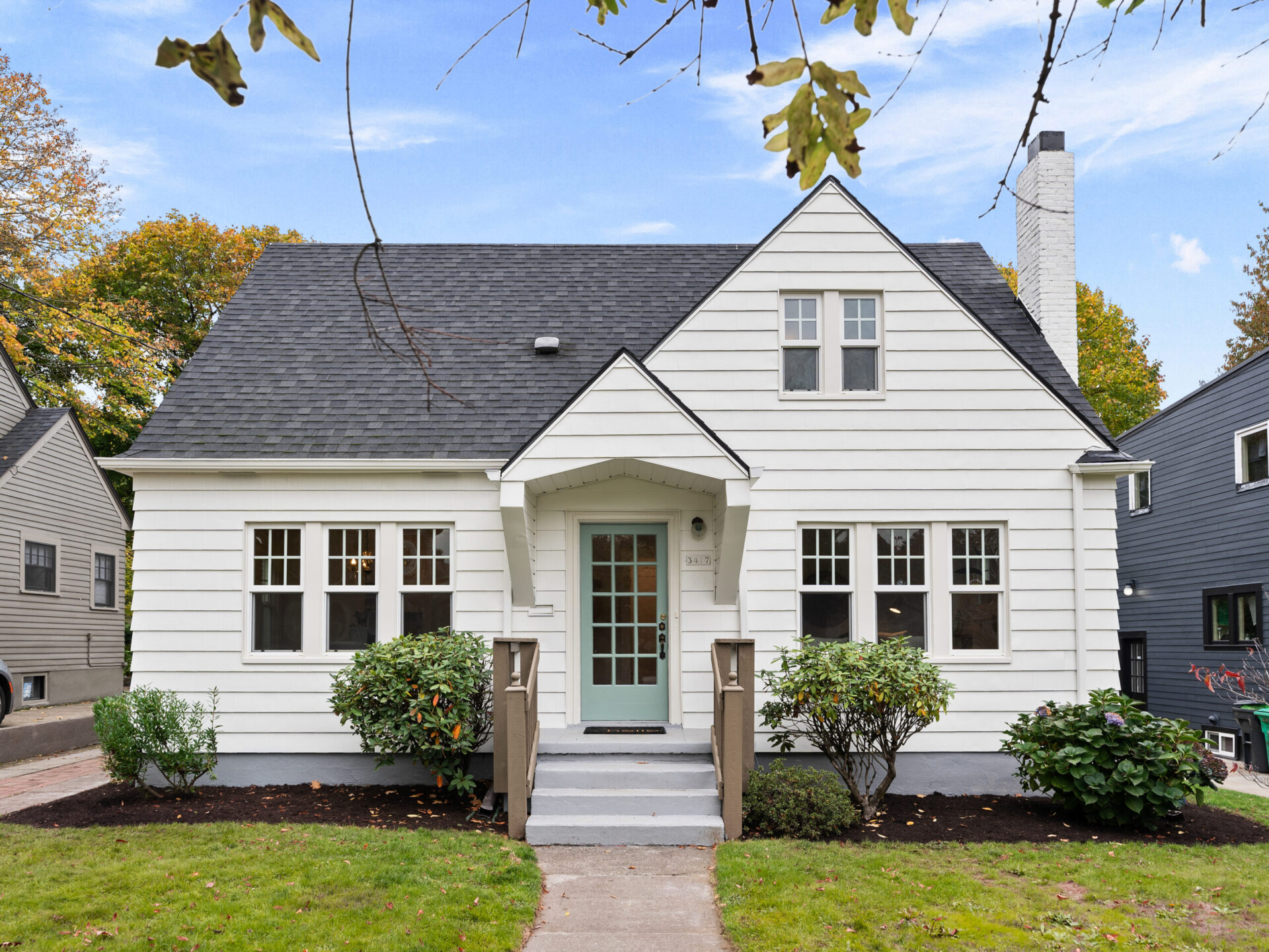 A charming white house with a steep, dark gray roof and chimney, featuring a mint green front door, surrounded by manicured shrubs. The sky is clear, and autumn leaves are visible on nearby trees and the ground—discover this gem with a top Portland Realtor today.