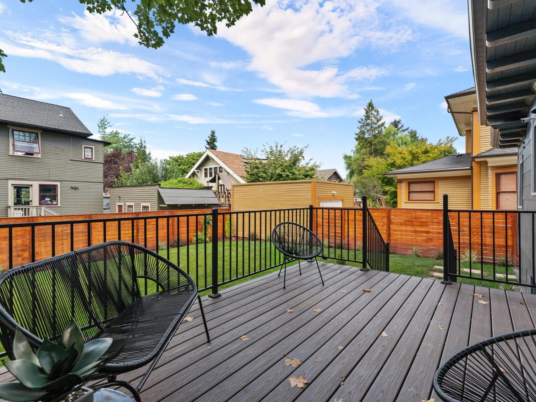 Outdoor deck with wire chairs and a potted plant, overlooking a grassy backyard in charming Portland, Oregon. A wooden fence surrounds the area, while the partly cloudy sky reveals glimpses of blue over neighboring houses.