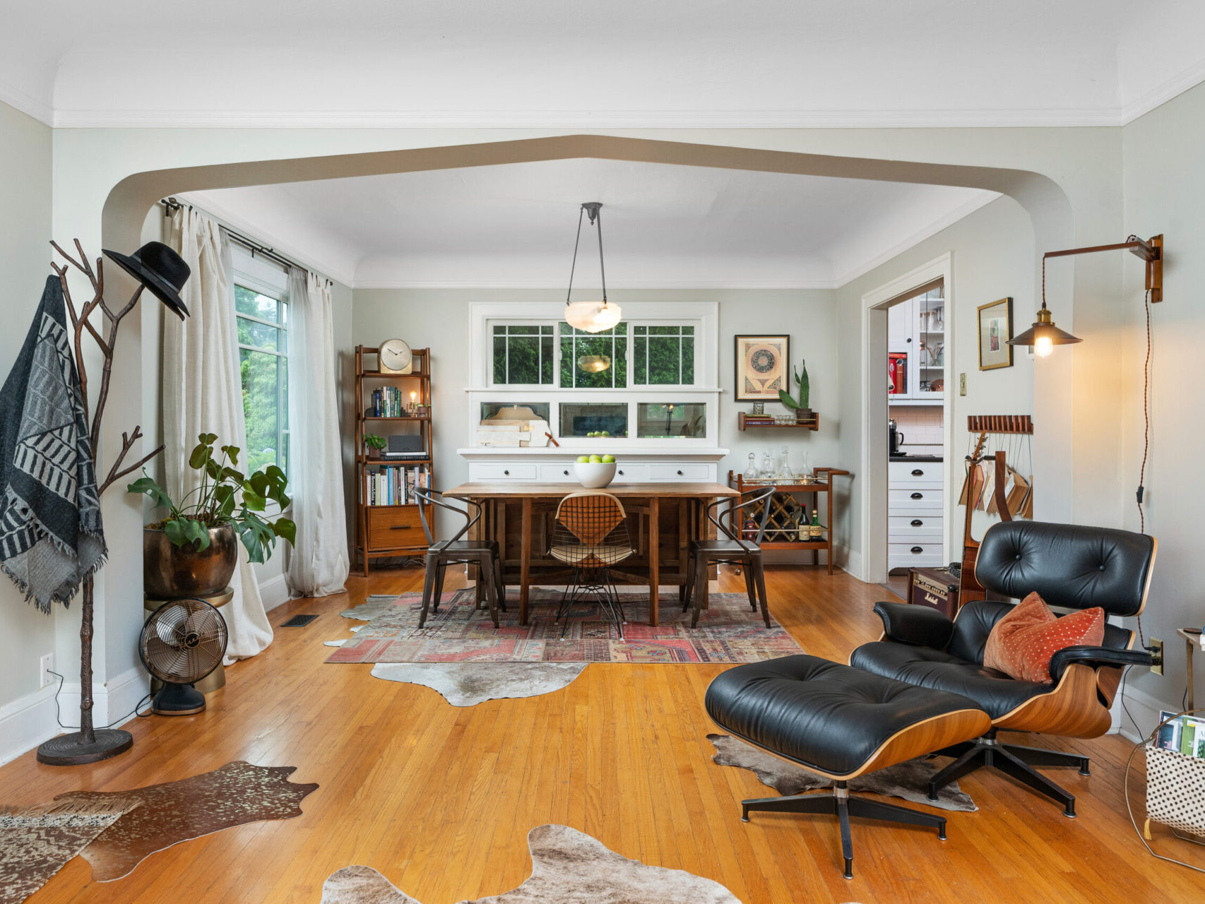 A cozy living room with a mid-century modern chair and footrest, a wooden dining table for six, and a shelf adorned with books and plants. Warm lighting and hardwood floors complete the Portland Oregon real estate gem, inviting you to explore more with your trusted Portland realtor.