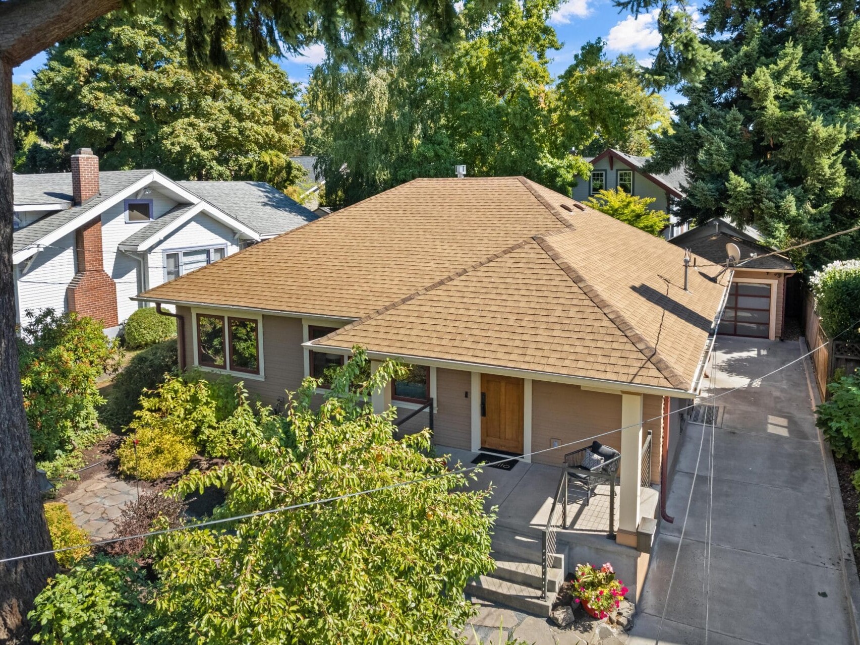 Aerial view of a tan house with a large sloped roof, nestled in the lush greenery typical of Portland, Oregon. A driveway leads to a detached garage. The house features a front porch and well-maintained garden. Other houses and a clear sky stretch across the Portland skyline in the background.