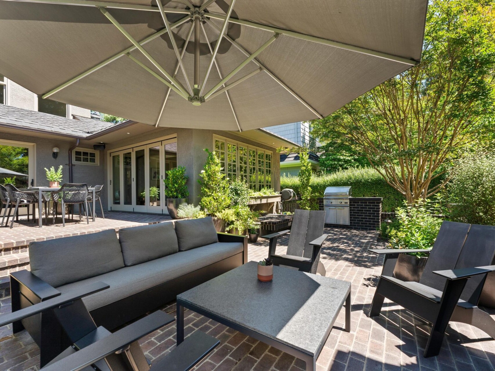 A modern outdoor patio in Portland, Oregon real estate features a large umbrella, gray cushioned sofa, and black chairs around a table. Brick flooring and lush greenery add to the inviting atmosphere. A dining table and grill are visible in the background, perfect for a Portland realtor showcase.