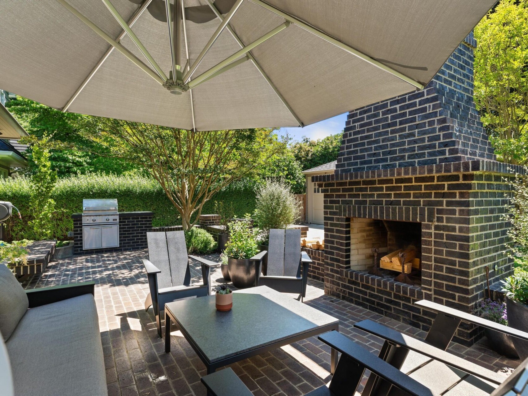 A patio with a large umbrella shades a table and chairs. A brick fireplace sits nearby, while a barbecue grill is in the background. Surrounded by lush greenery, this cozy outdoor living space epitomizes Portland Oregon real estate at its finest.
