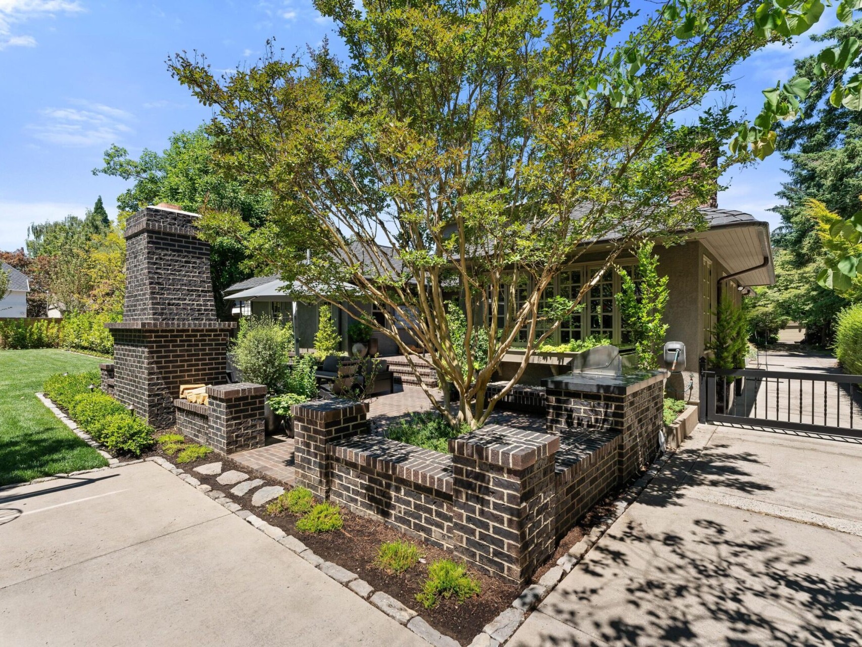 A charming outdoor patio with a brick fireplace and grill, surrounded by lush Portland greenery. The intricate brickwork creates a cozy corner, ideal for any Portland realtor's portfolio. A paved pathway and small lawn border the area, while trees provide shade to enhance the tranquil atmosphere.