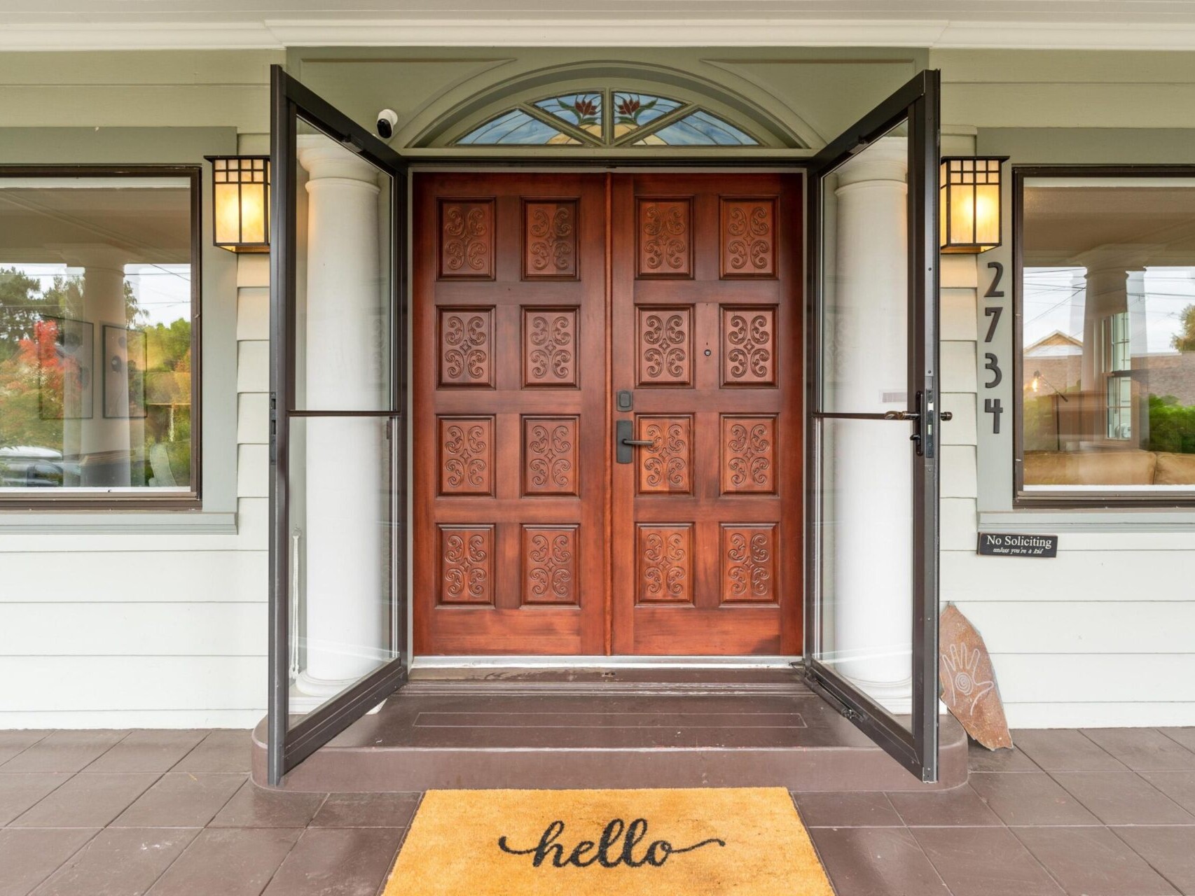 A wooden front door with intricate carvings is flanked by two white columns. A welcome mat with hello is placed on the porch floor. The house number 2734 is displayed on the siding next to the door.