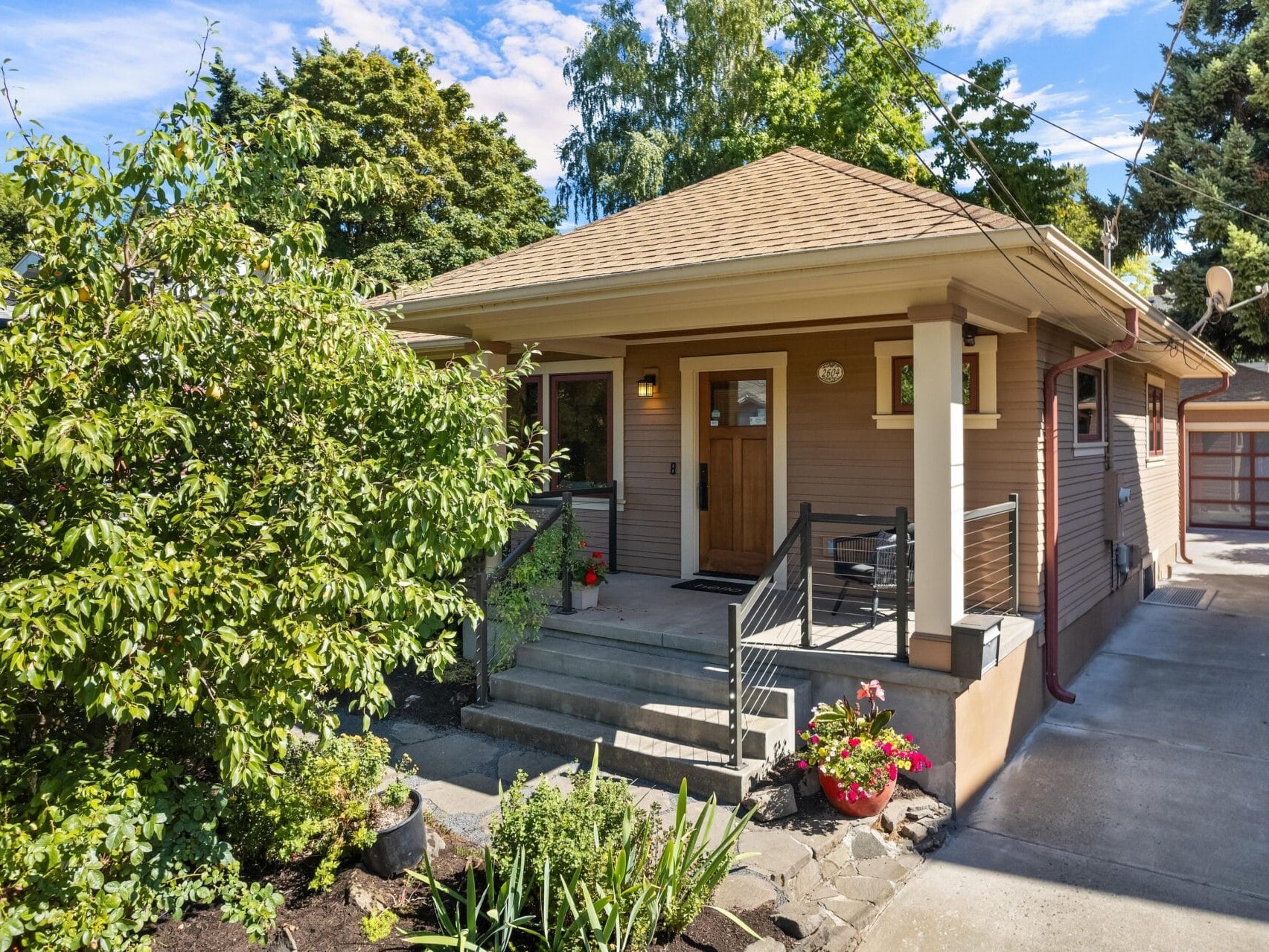 A charming single-story house in Portland, Oregon, features a covered front porch, wooden door, and small garden. Concrete steps lead to the entrance, flanked by potted plants. A driveway extends to a detached garage in the background amid lush greenery.