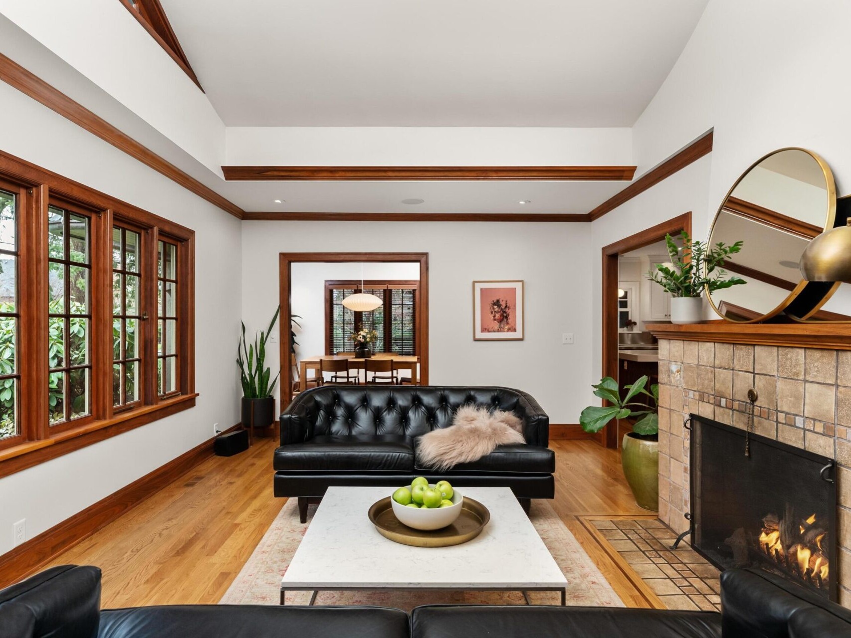 A cozy living room in a Portland Oregon real estate gem features a black leather sofa with a fur throw, a fireplace ablaze, and a coffee table adorned with green apples. Large wooden windows and a mirror above the mantel enhance the space, while indoor plants offer lush greenery.