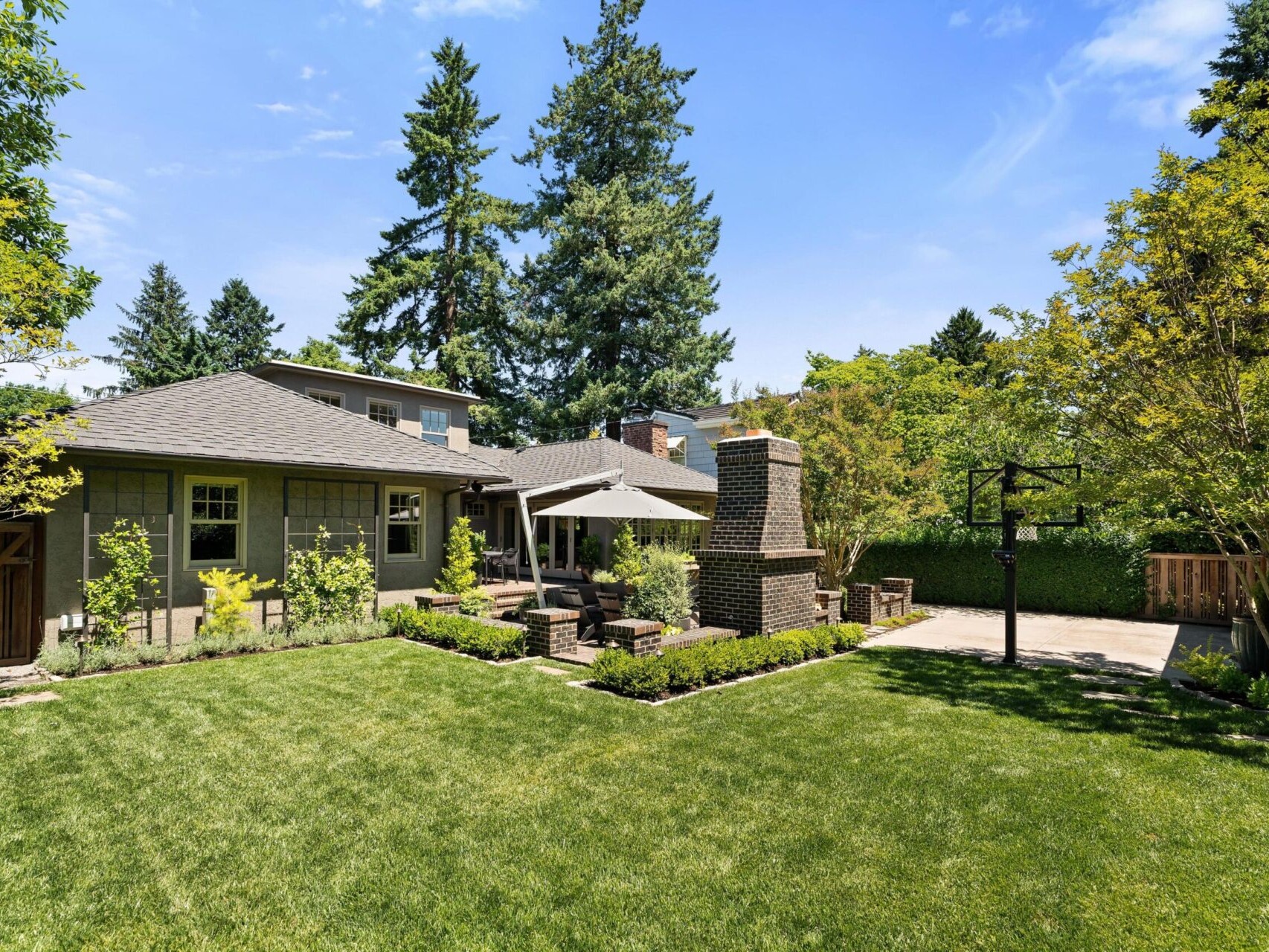 A serene backyard in the heart of Portland Oregon real estate features a well-maintained lawn and patio area with outdoor seating and a large umbrella. Tall trees and manicured shrubs border the space, while a brick chimney and basketball hoop add functionality and charm.
