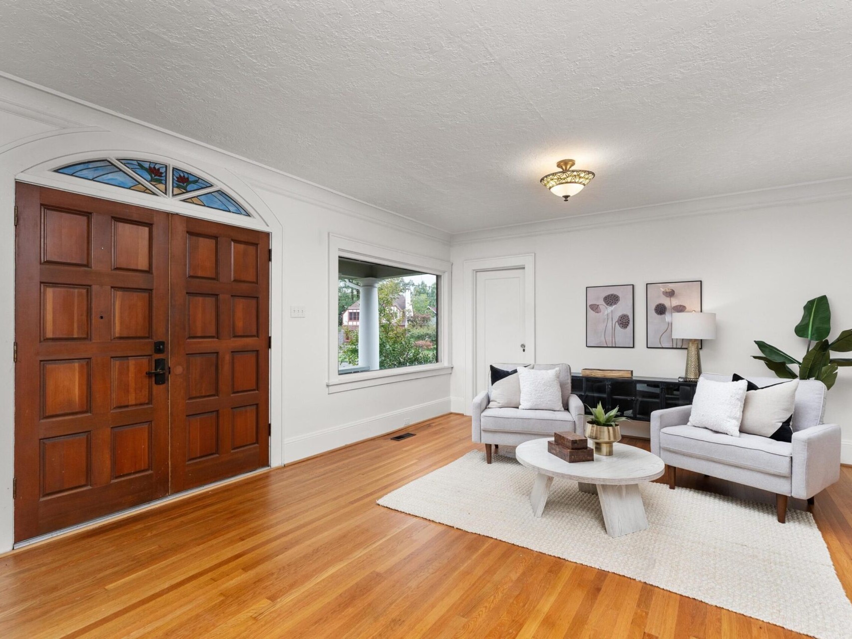 A cozy living room with two gray armchairs, a round coffee table, and a large window. The space features wooden floors, a patterned ceiling, and a pair of decorative prints on the wall. A wooden double door is shown on the left.