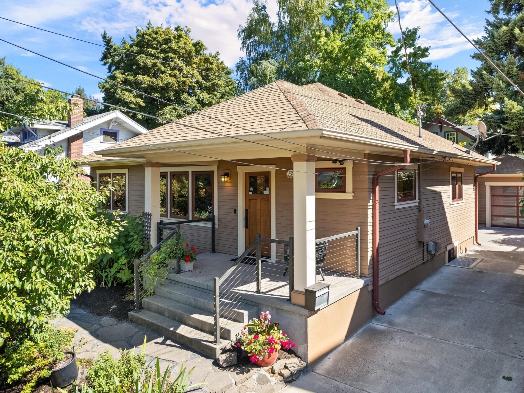 A charming one-story house in Portland, Oregon, with a sloped roof and beige siding. It features a covered front porch with steps adorned by potted plants. The driveway leads to a detached garage, and the yard is lush with green trees and shrubs under a blue sky.