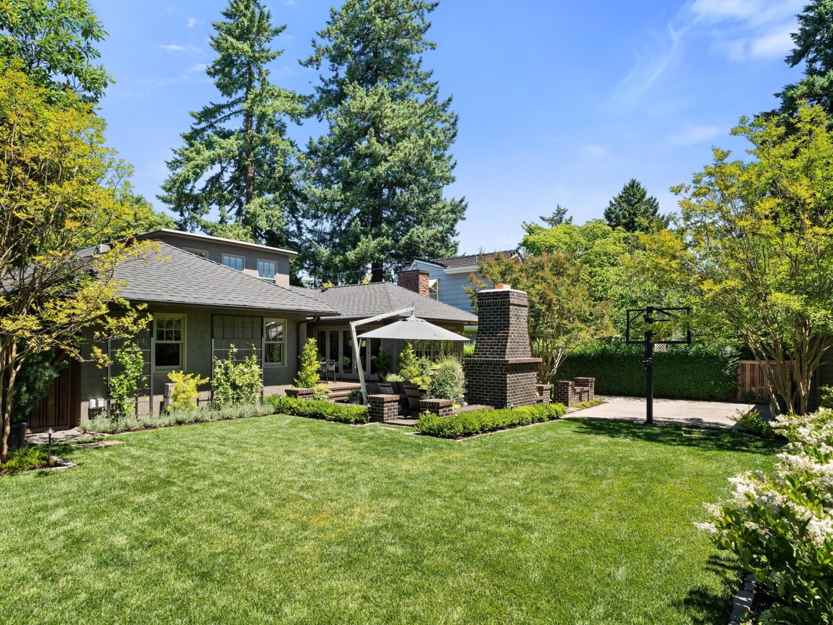 A backyard oasis in Portland Oregon Real Estate, this space features a lush green lawn, shaded patio with outdoor furniture, brick fireplace, and a basketball hoop. Tall trees and manicured bushes surround the area, with a house and shed under a clear blue sky.