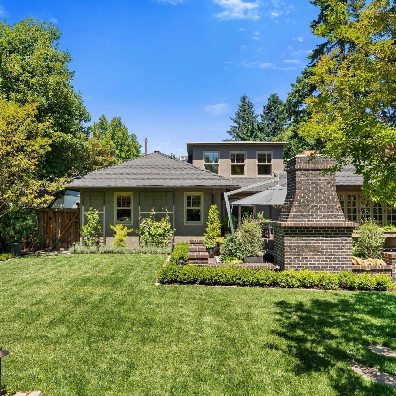 A modern two-story house with a brick chimney is nestled within Portland Oregon Real Estate, surrounded by a lush green lawn with various trees and shrubs. In the backyard, neatly trimmed hedges and a stone pathway lead to the patio area, all under a clear blue sky.