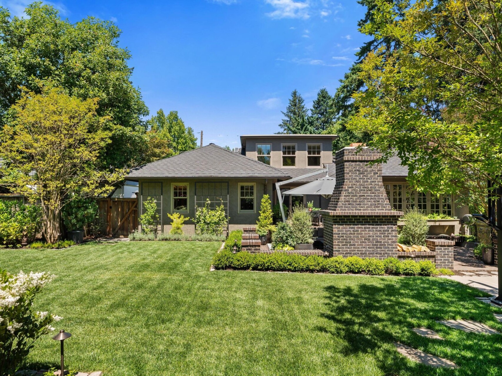 A modern two-story house with a brick chimney is nestled within Portland Oregon Real Estate, surrounded by a lush green lawn with various trees and shrubs. In the backyard, neatly trimmed hedges and a stone pathway lead to the patio area, all under a clear blue sky.