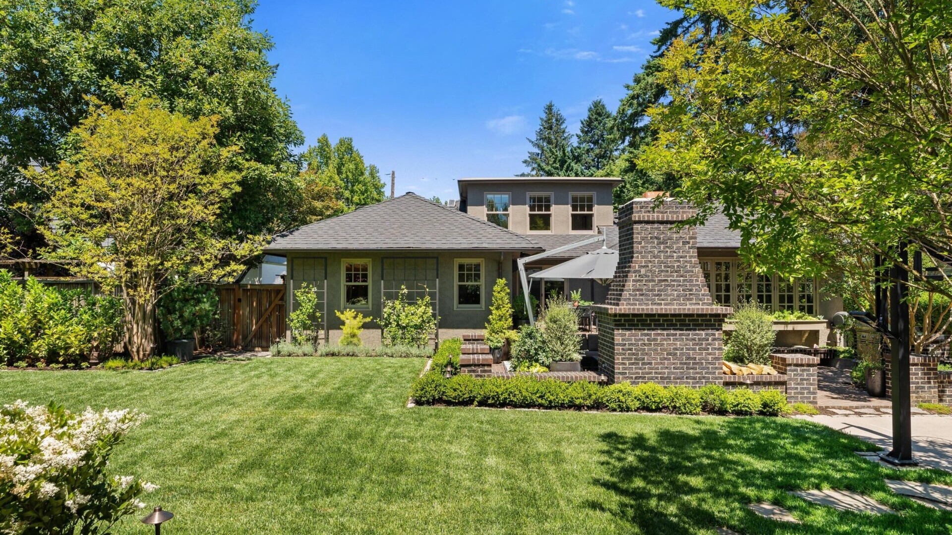 A modern two-story house with a brick chimney is nestled within Portland Oregon Real Estate, surrounded by a lush green lawn with various trees and shrubs. In the backyard, neatly trimmed hedges and a stone pathway lead to the patio area, all under a clear blue sky.