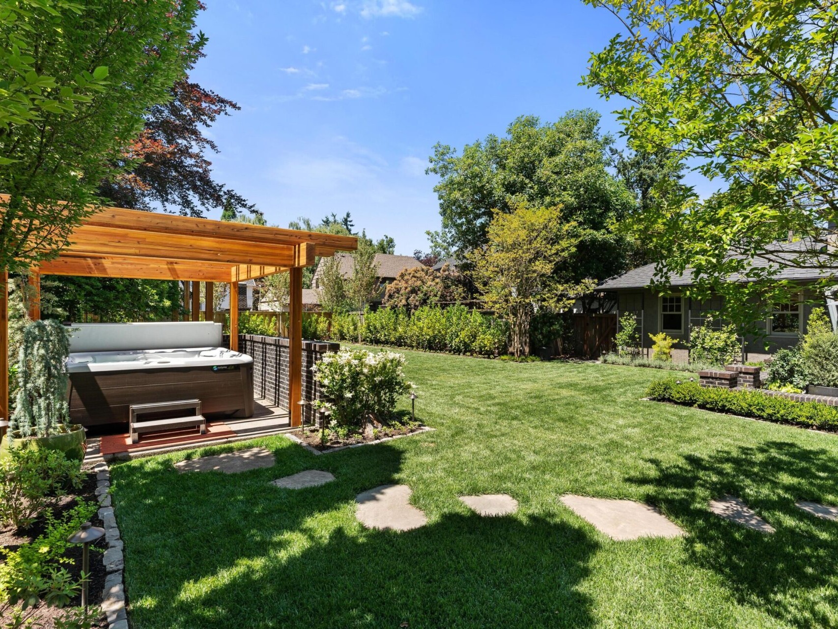 The serene backyard boasts a lush green lawn, bordered by trees and shrubs. A wooden pergola shelters a hot tub to the left, while stone stepping paths meander through the inviting space. Ideal for Portland Oregon real estate seekers, this oasis complements the brickwork house under clear, sunny skies.
