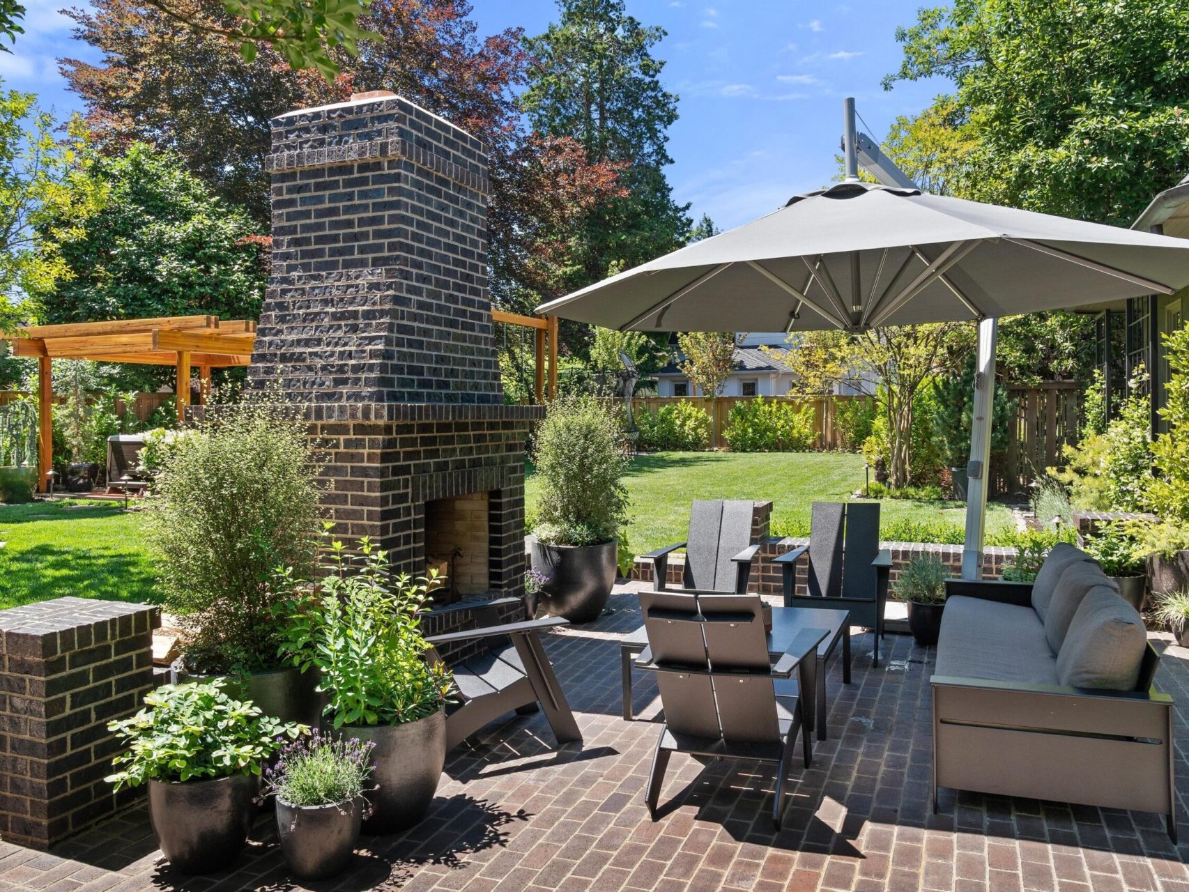 A backyard patio featuring a brick fireplace, modern outdoor furniture, and a large umbrella is perfect for enjoying Portland Oregon real estate. Potted plants and trees decorate the area, while a pergola and well-maintained lawn enhance the charm on this bright, sunny day with clear skies.