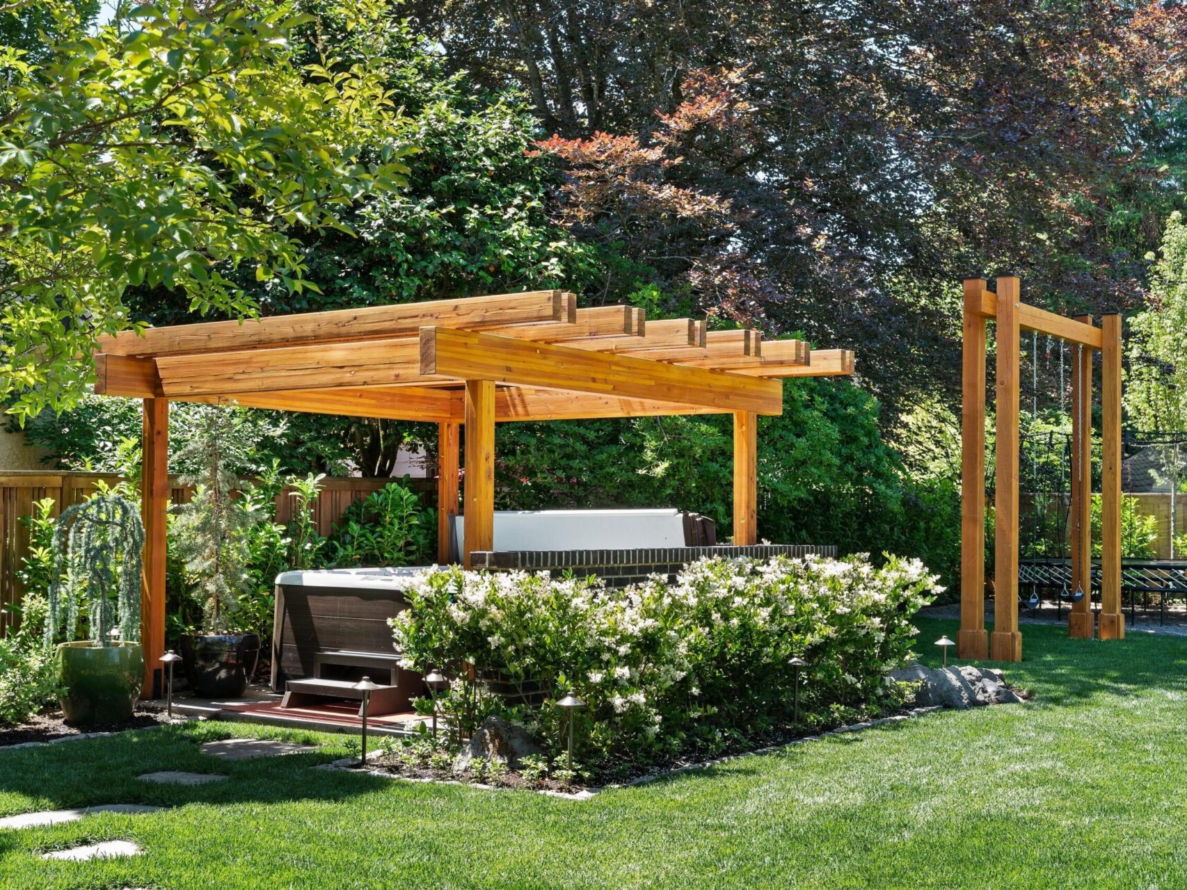 A backyard with a wooden pergola over a hot tub, surrounded by lush green plants and blooming white flowers. Nestled within Portland's serene landscapes, the area features neatly manicured grass and is framed by trees, offering a peaceful retreat that any top Portland Realtor would highlight.