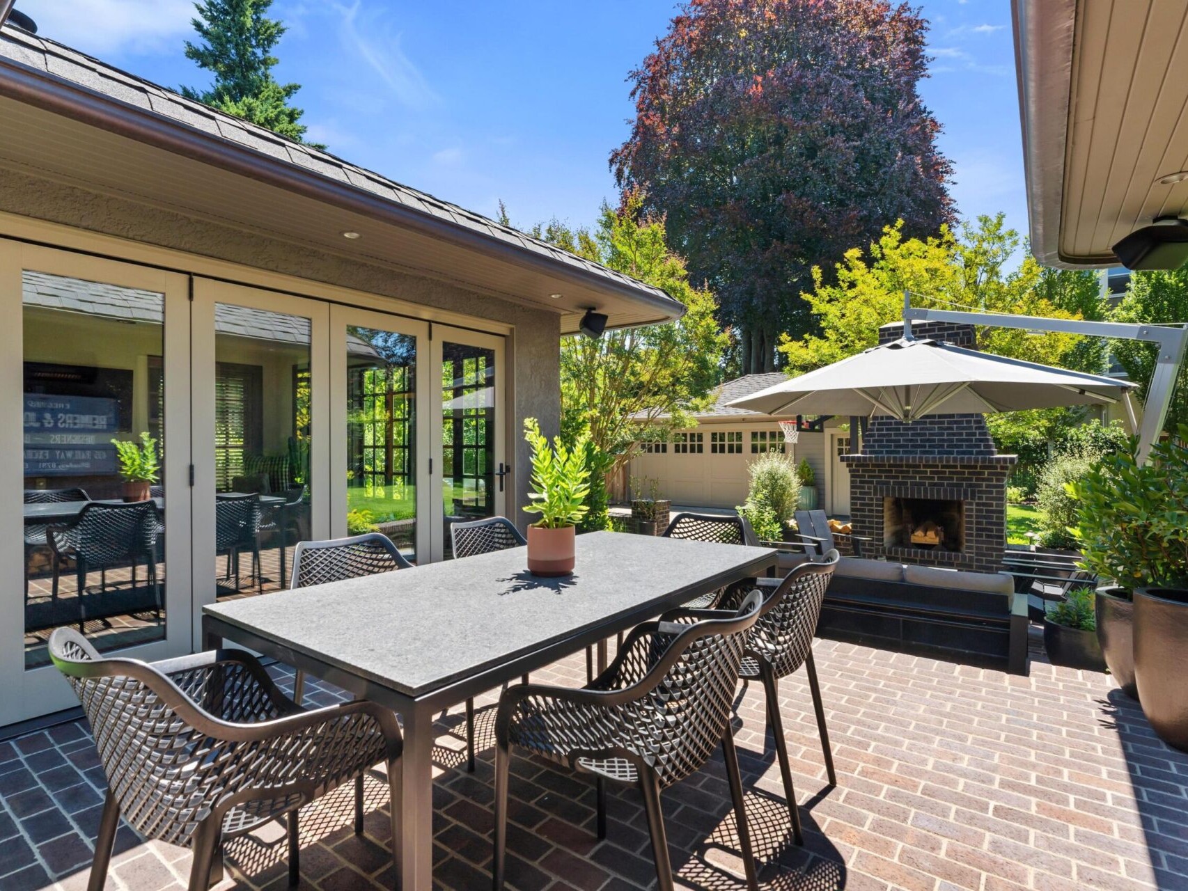 Outdoor patio with a large dining table and eight chairs on a brick floor. A fireplace with a chimney is nearby, and a large umbrella provides shade. Surrounding trees and greenery enhance the tranquil atmosphere, reflecting the charm Portland Oregon Real Estate offers.