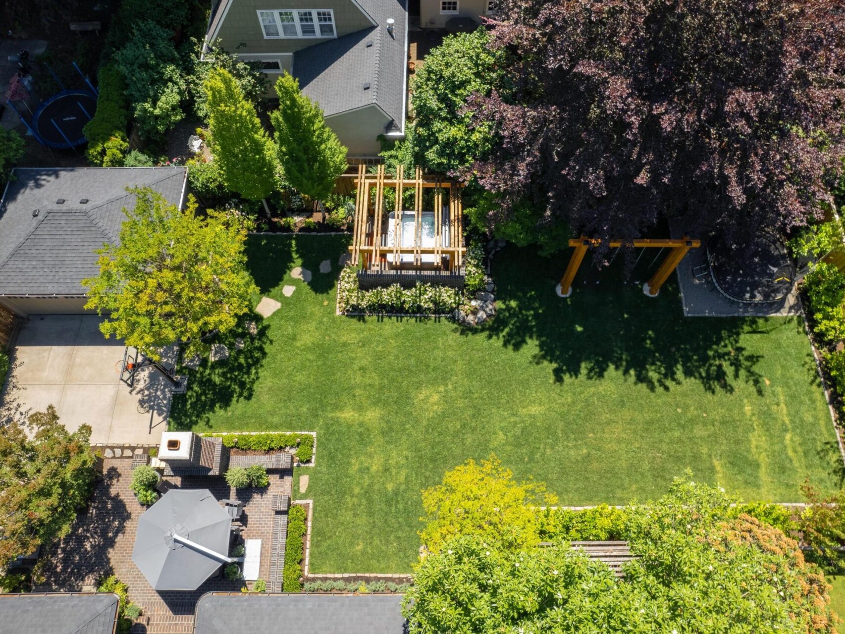 Aerial view of a landscaped Portland Oregon real estate gem, featuring a green lawn, pergola, and two gray rooftops. There's a paved patio with an umbrella on the left, and the lawn is bordered by trees and shrubs—perfectly showcased by a top real estate agent.