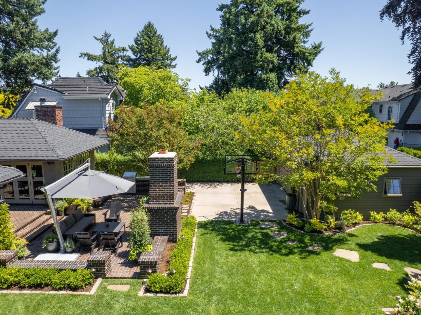Aerial view of a charming Portland Oregon real estate gem: a backyard with a patio featuring a dining set under an umbrella, beside a brick chimney. It boasts a grassy lawn, basketball hoop on paved area, and lush trees and shrubs, with nearby houses completing the serene scene.
